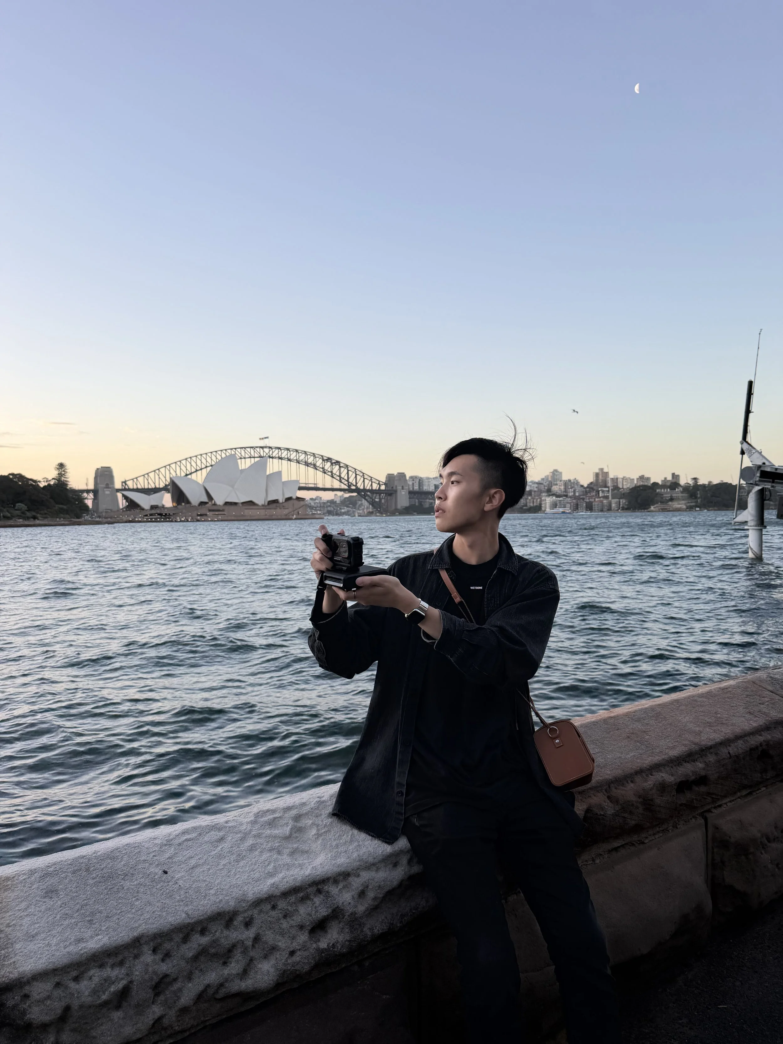 A young man standing by the water's edge, holding a camera, with the Sydney Opera House and Harbour Bridge in the background during sunset.