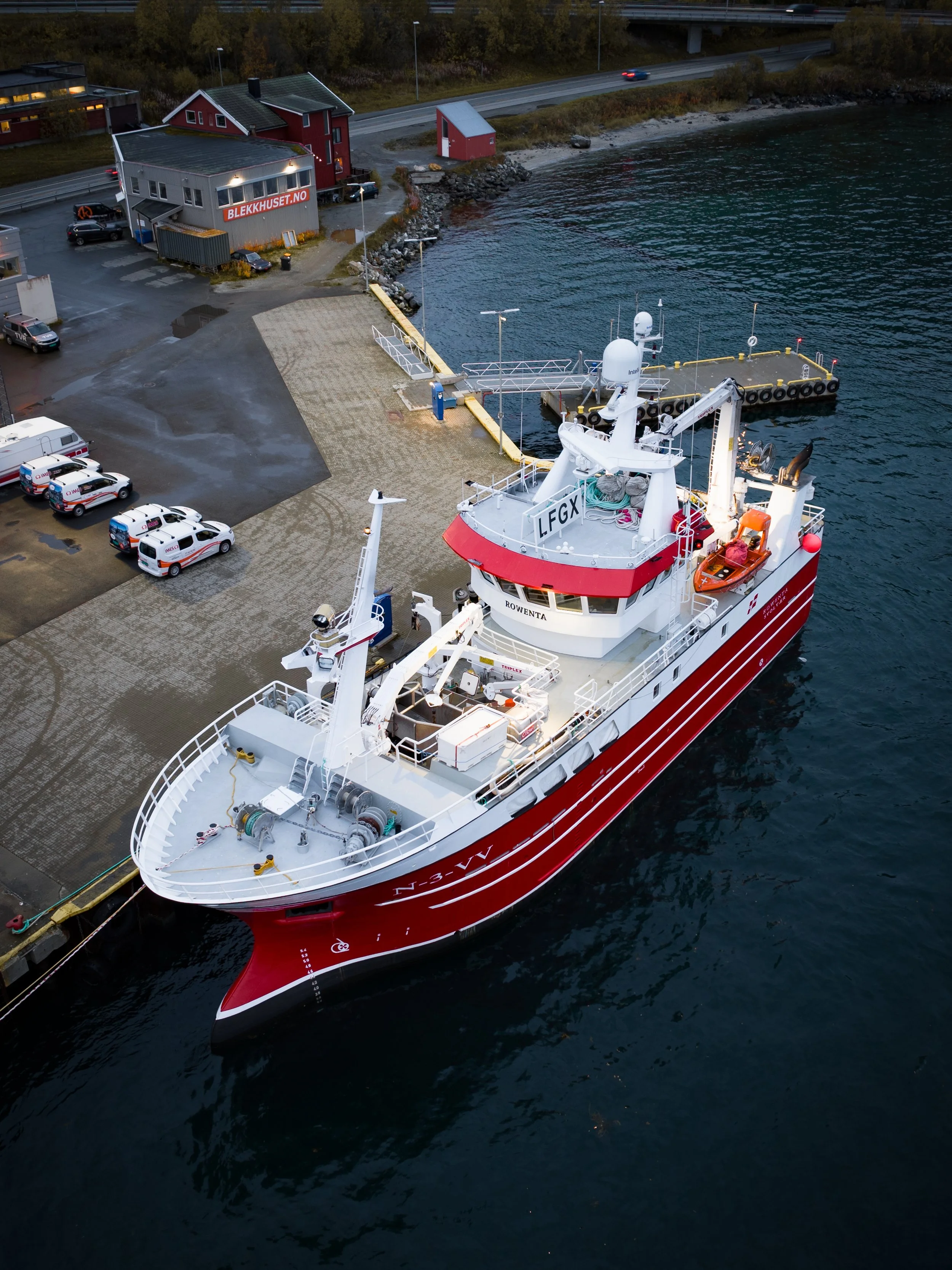 A large white and red research or supply vessel docked at a harbor with a concrete pavement, several parked cars, and a few small red buildings nearby. The water is calm and dark, with some ripples, and there are trees and a road in the background.