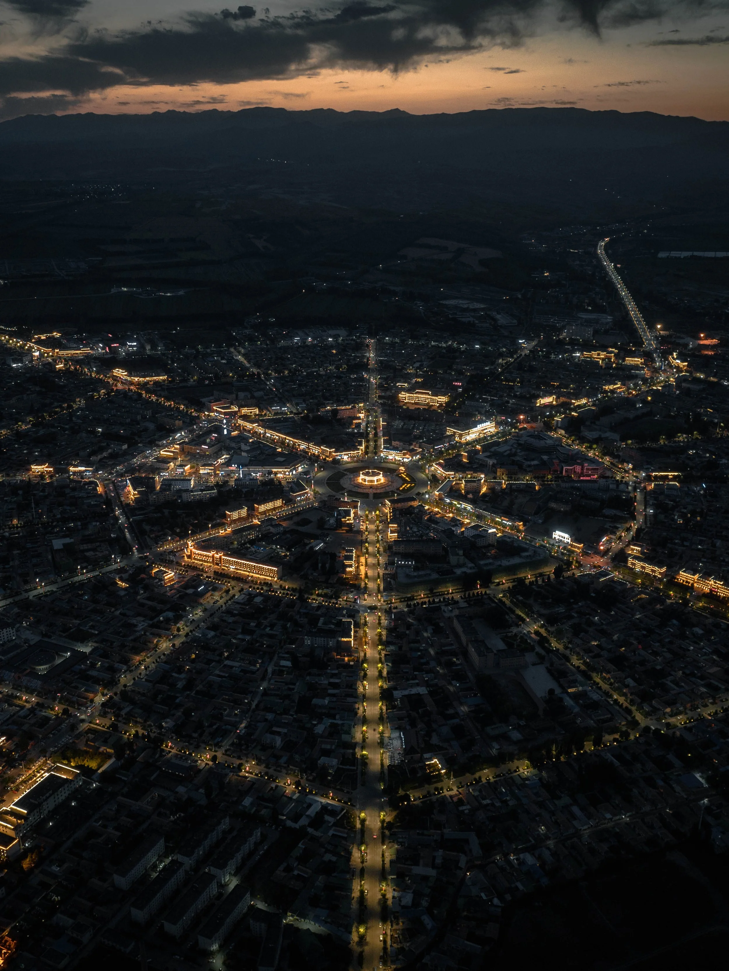 Aerial night view of a city with illuminated streets and buildings, mountains in the background, and a dark cloudy sky.
