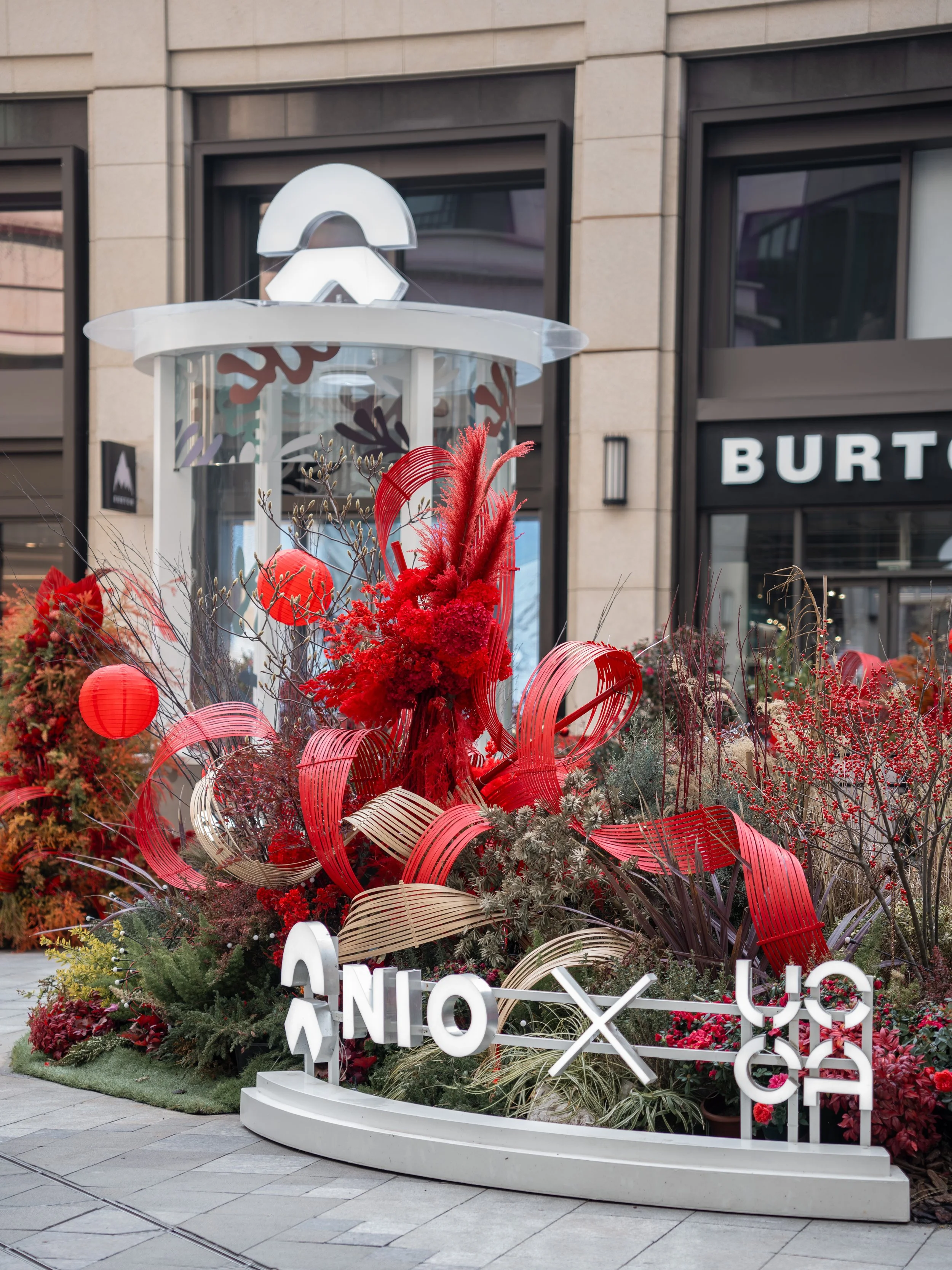 Public outdoor display featuring red and white decorative elements and flowers, with large white letters and geometric structures, in front of a retail building with dark-framed windows and a sign reading 'BURT'.