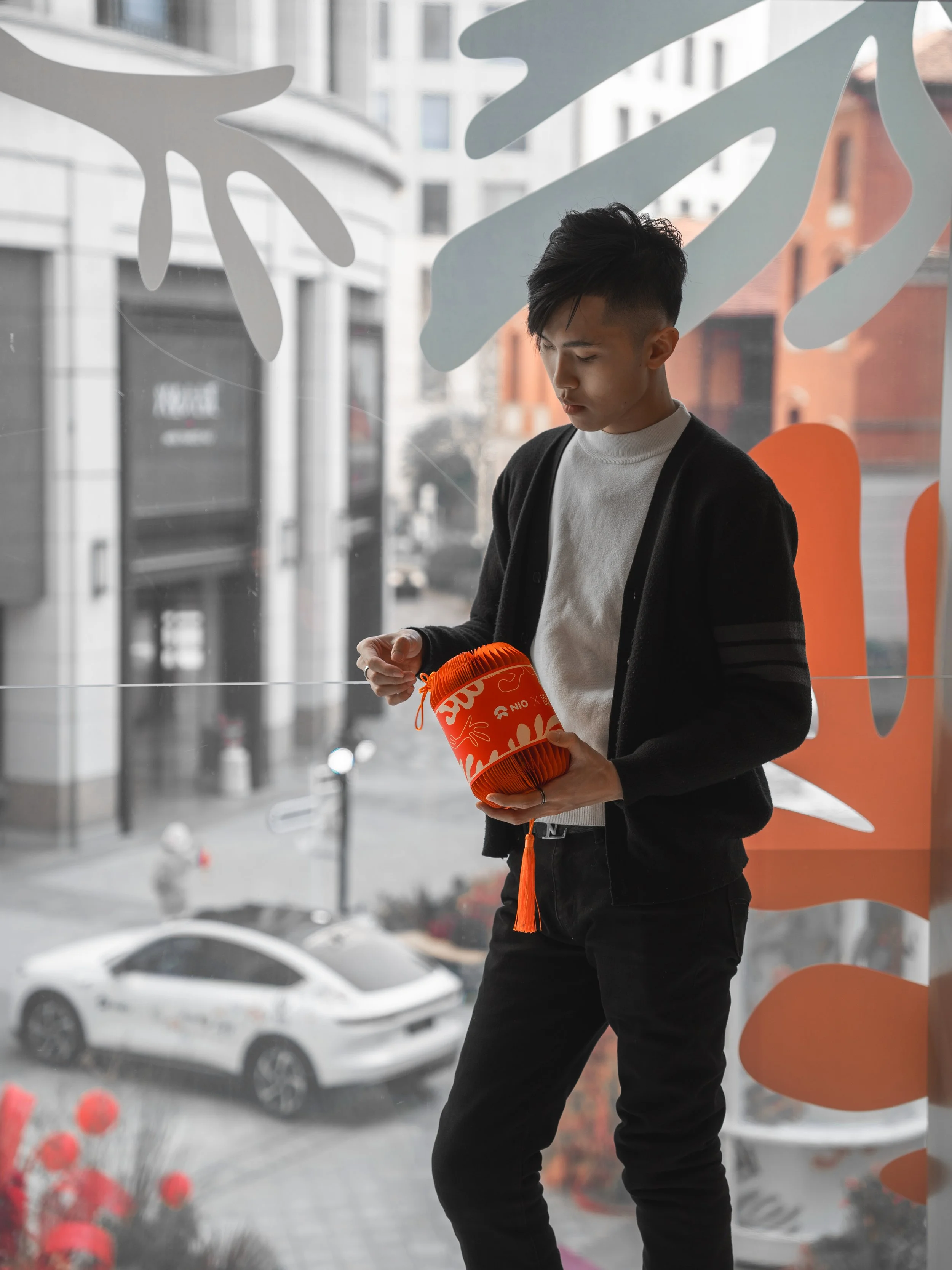 Young man with black hair, wearing a black jacket over a white shirt, holding an orange paper lantern in front of a large glass window with city buildings and a white car outside.