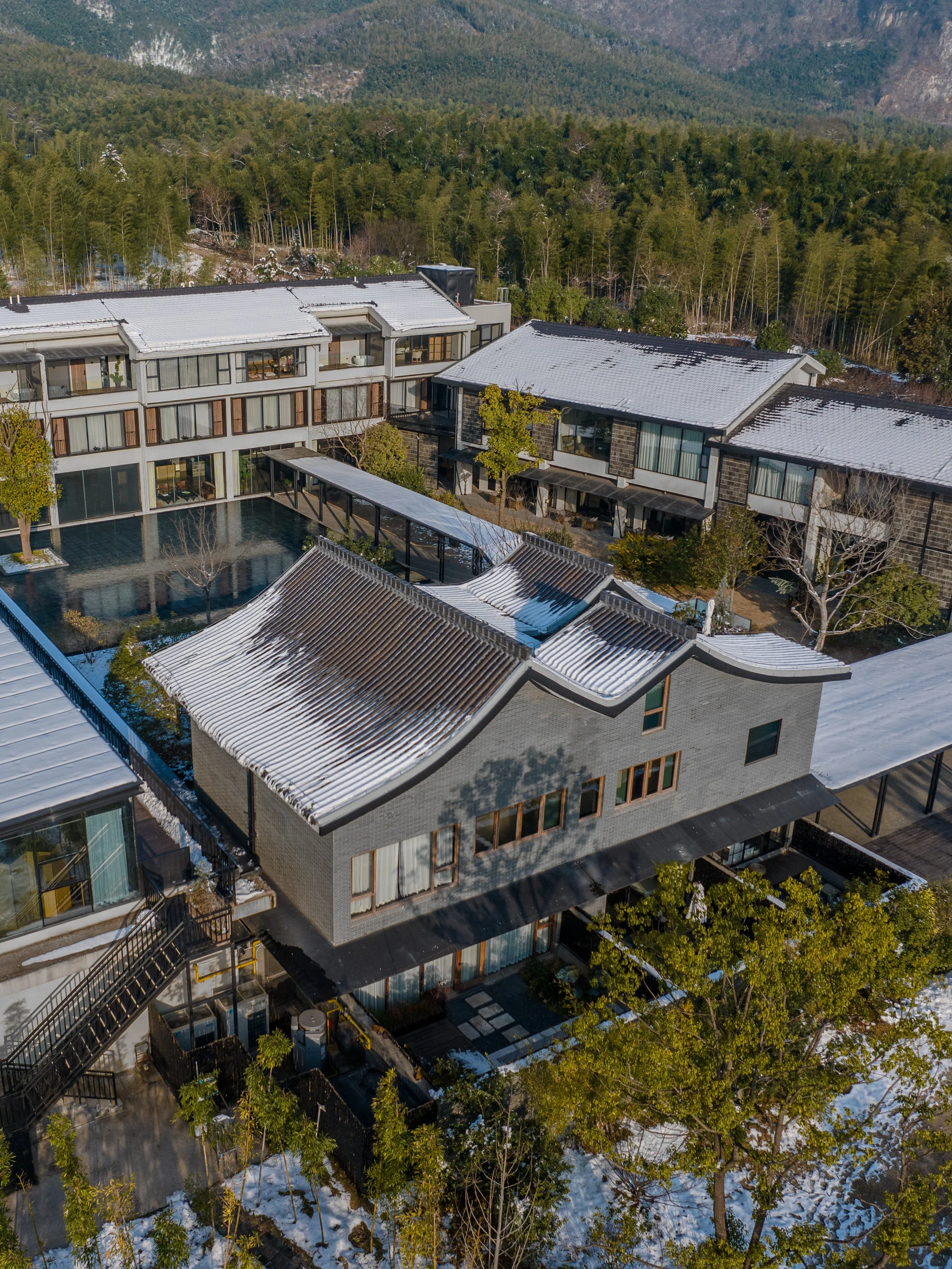 An aerial view of a modern residential building surrounded by trees and snow, with mountain forest in the background.