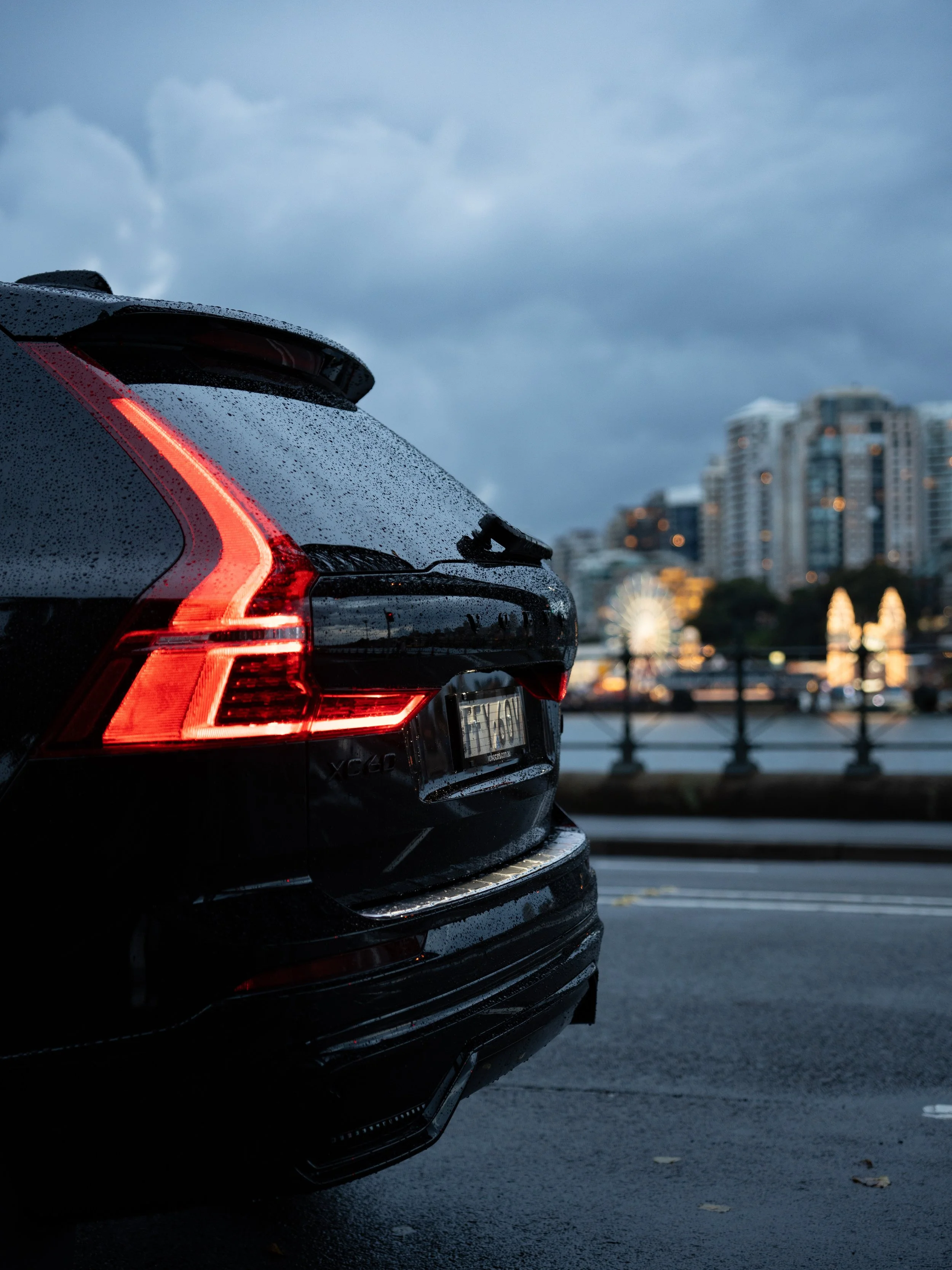 Close-up of the rear of a black car with taillights illuminated, parked on a wet street with city buildings and a cloudy sky in the background.