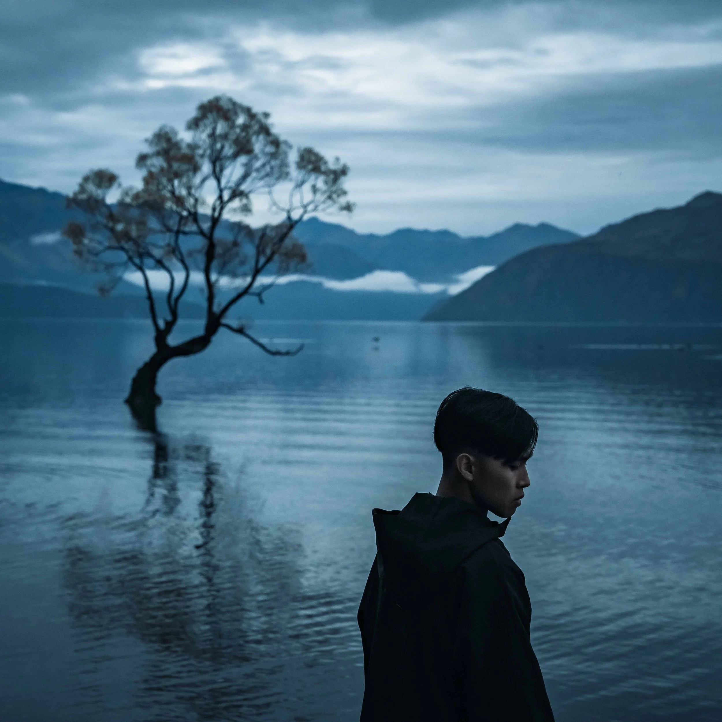 A silhouette of a person with short hair standing near a body of water during dawn or dusk, with mountains and a cloudy sky in the background.