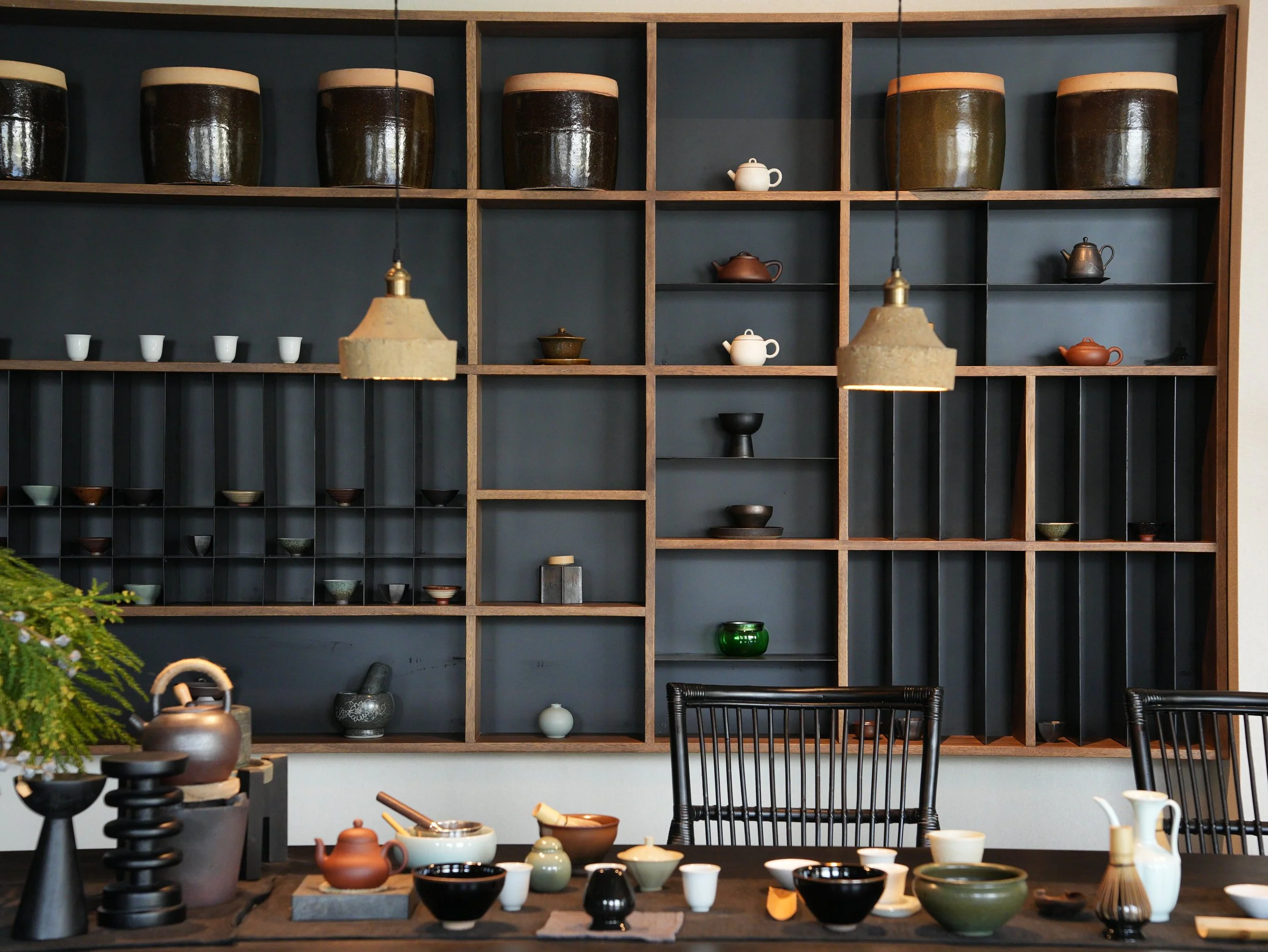 Display of various ceramic teapots and bowls on a table with a dark wooden shelving unit in the background, some teapots are brown, white, black, and orange, with a variety of shapes and sizes, and hanging pendant lights above.