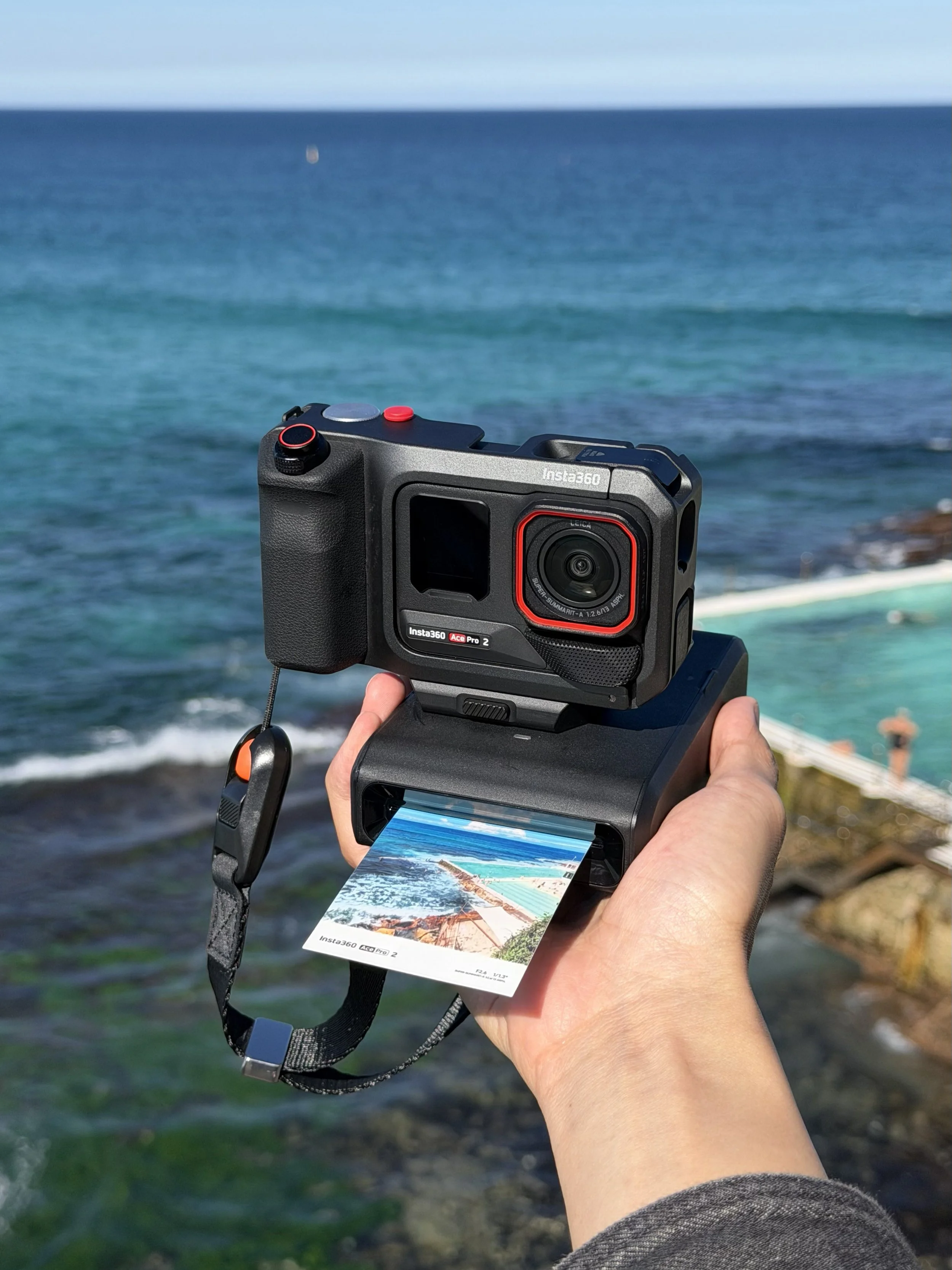 A person holding a camera and a printed photo of a beach scene with blue water and rocks, near the ocean with waves.