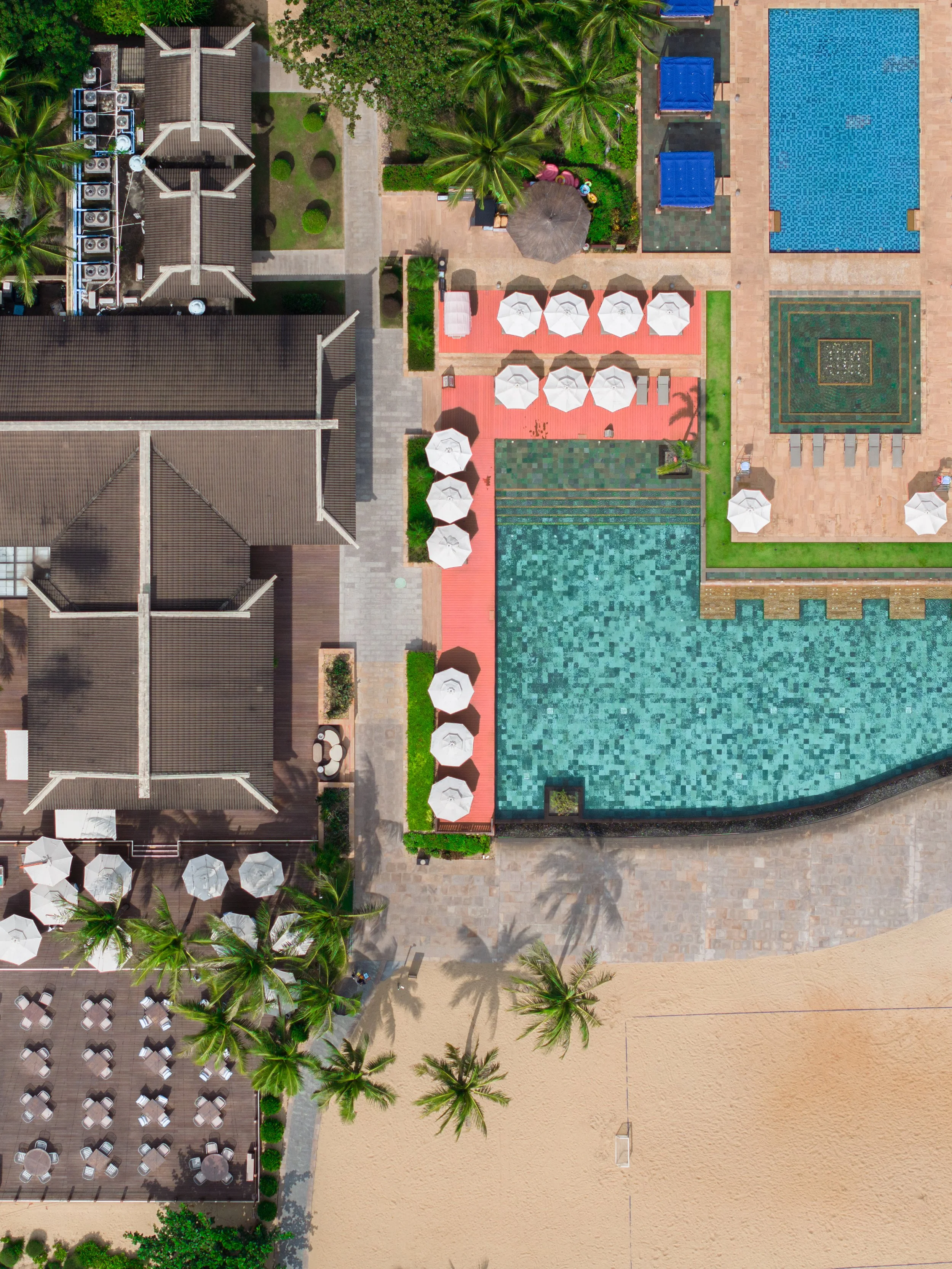 An aerial view of a resort or hotel pool area with lounge chairs, umbrellas, palm trees, a sandy area, and surrounding buildings.