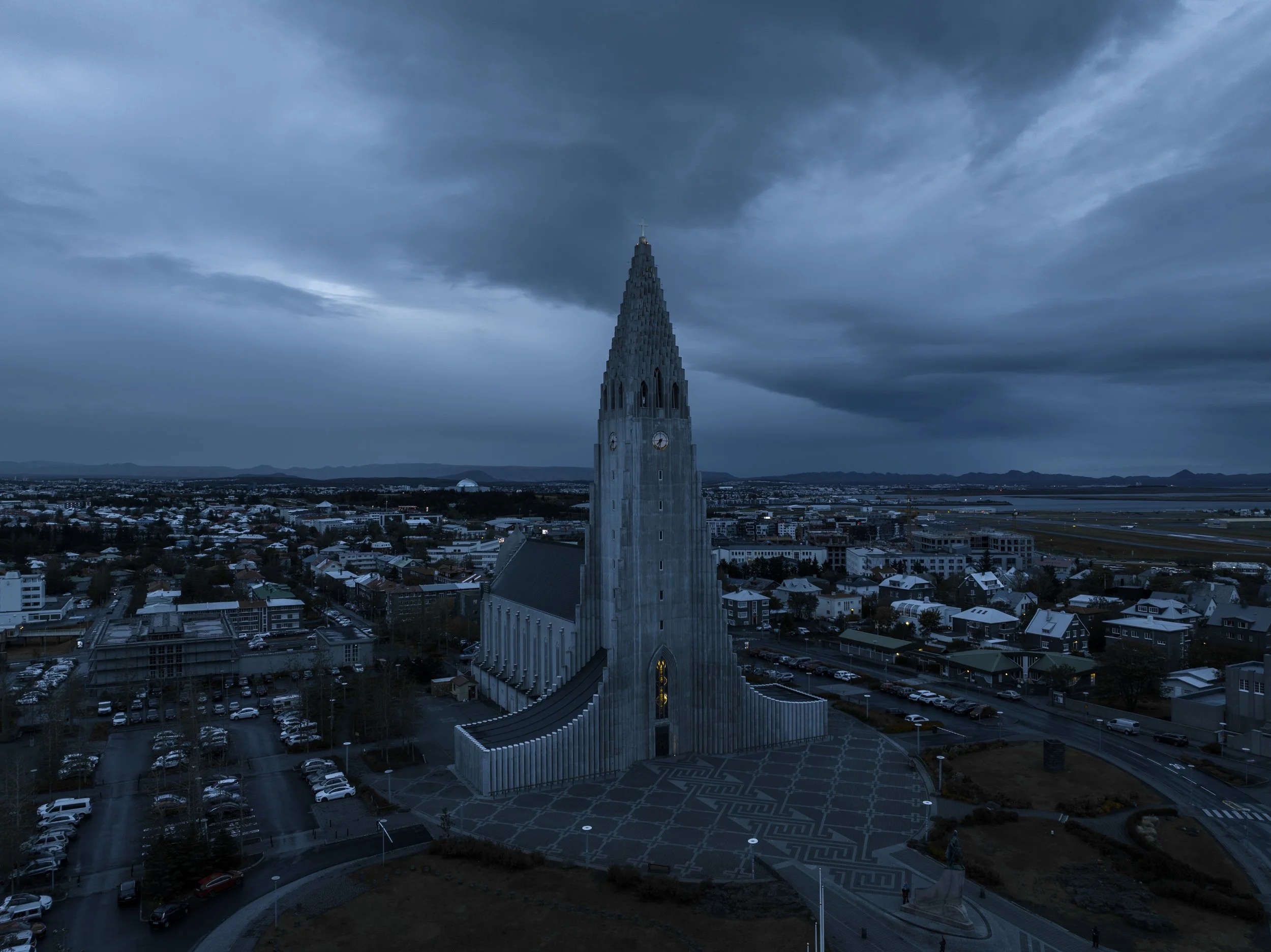 A tall church with a dark, pointed spire topped with a cross, standing over a cityscape on a cloudy, overcast day. The church has a modern, sleek design with narrow windows and a staircase at its base. The city below includes residential and commerci