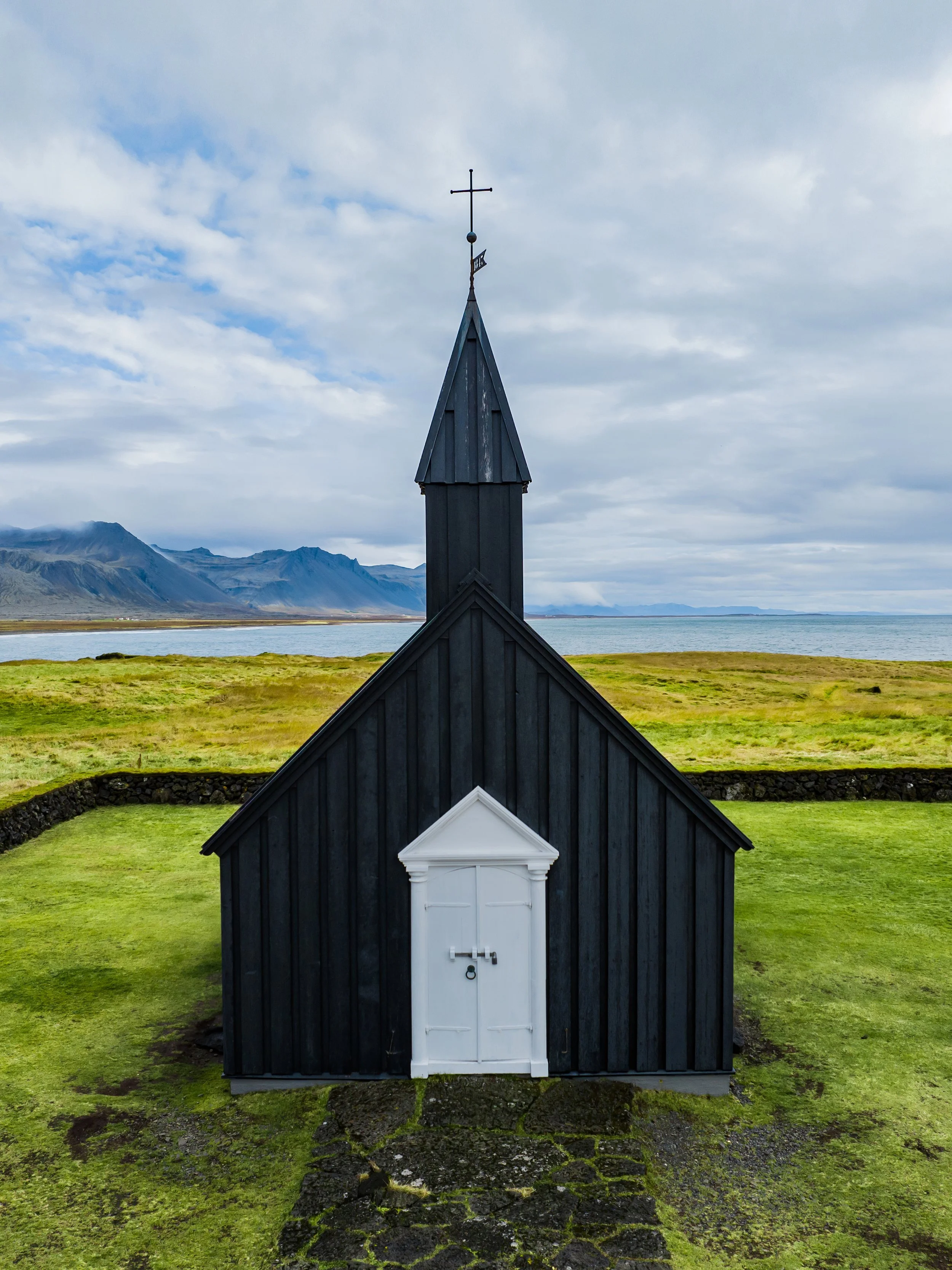 A small black wooden church with a white door, located in a grassy landscape with mountains and water in the background, under a cloudy sky.