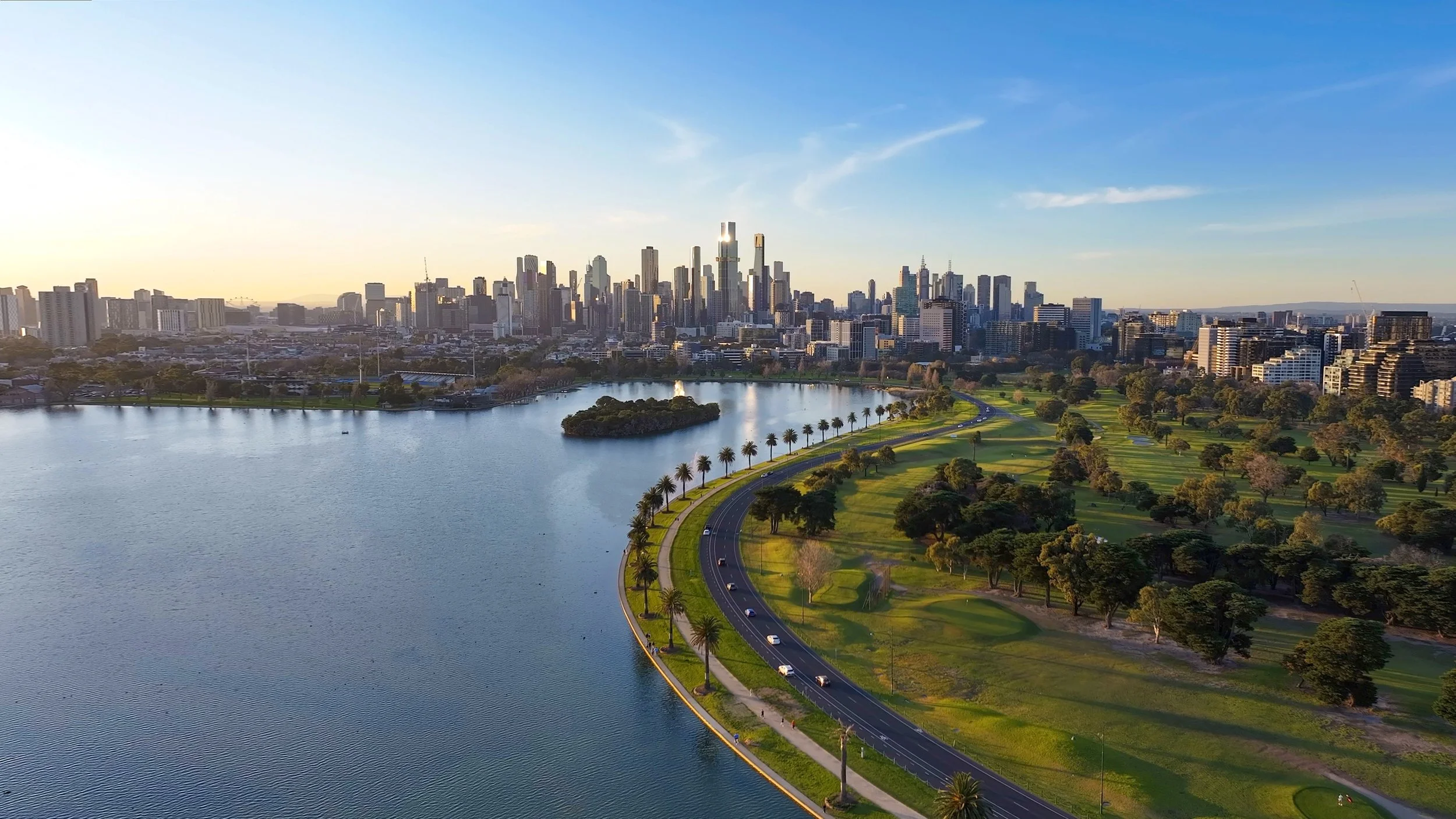 A city skyline with tall skyscrapers across a body of water. A green park with a curved road and palm trees runs beside the water in the foreground, while the skyline is illuminated by the setting sun.