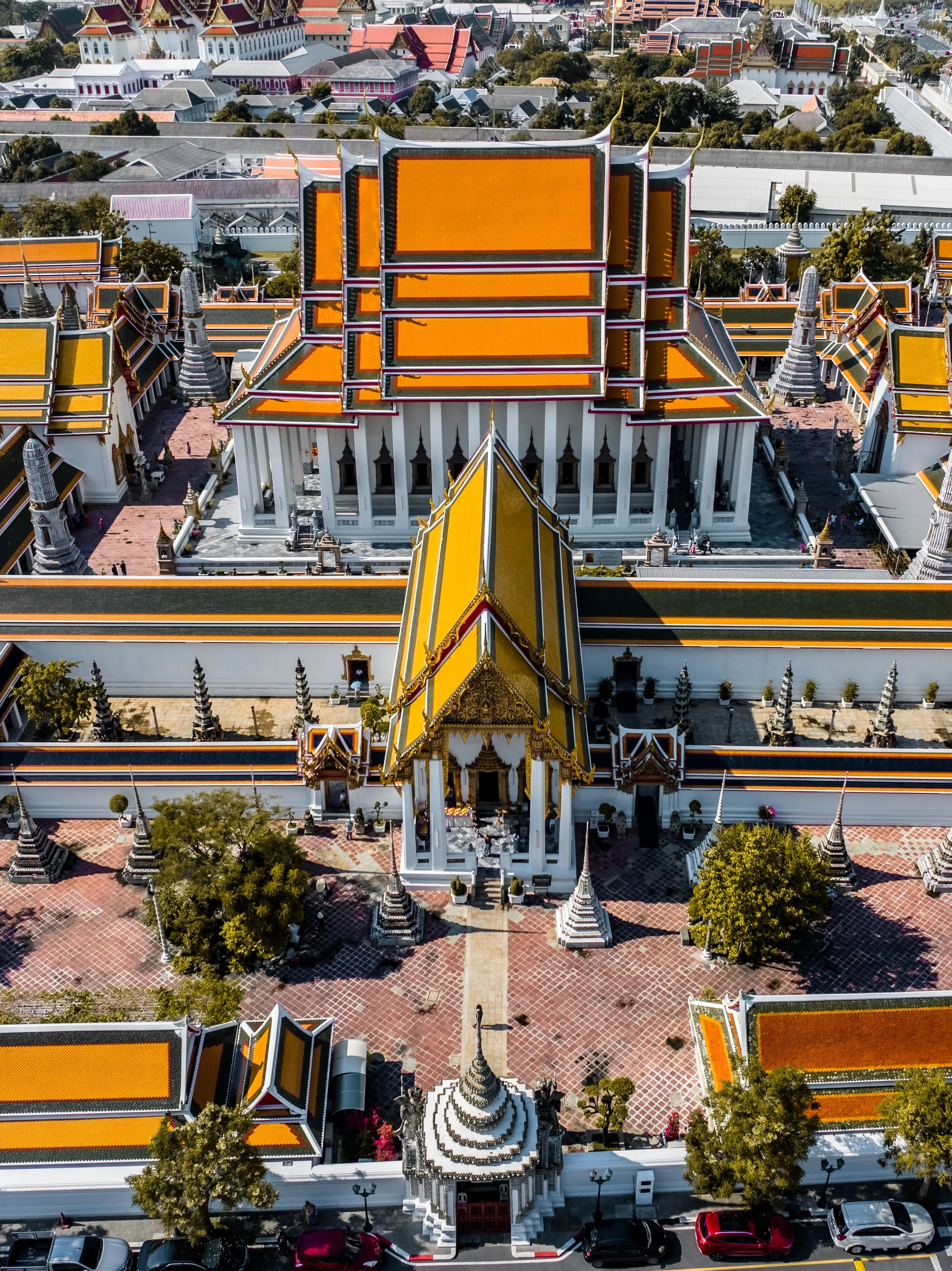 Aerial view of a traditional Thai temple complex with white walls, golden roofs, and decorative spires, surrounded by trees and parked cars.