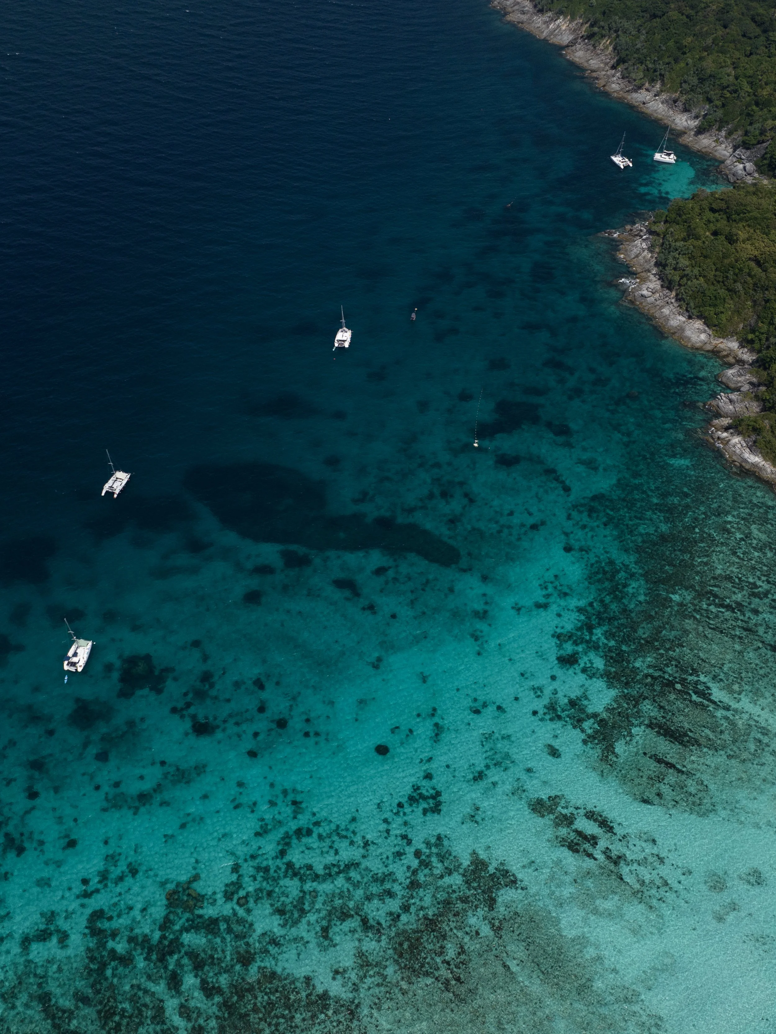 An aerial view of a coastline with clear blue water and several sailboats anchored near the rocky shore, surrounded by lush green trees.