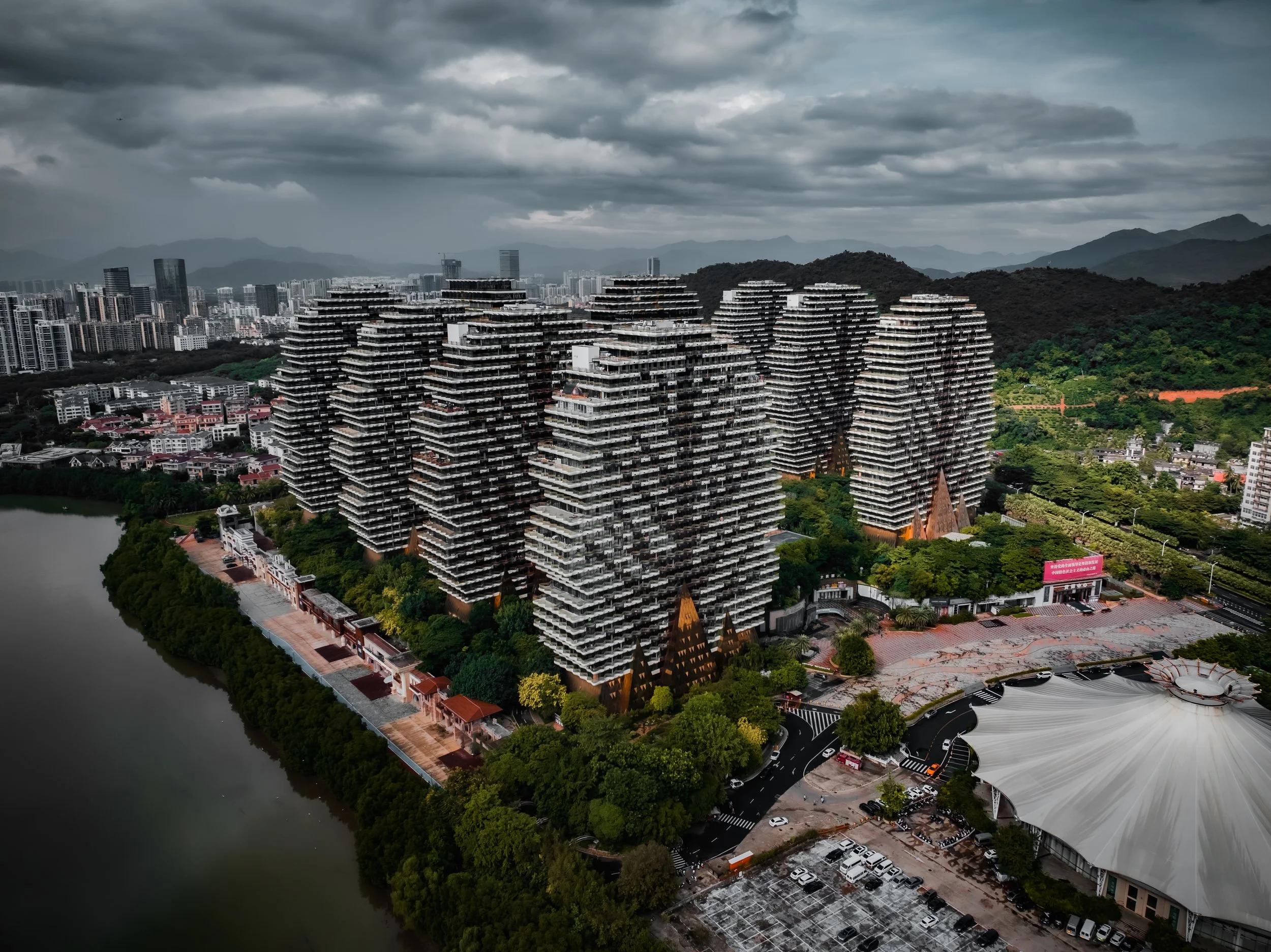 A cityscape with high-rise buildings, mountains in the background, and dark clouds overhead. The image shows a river on the left side, and a park or green area with trees near the buildings. There is also a white tent-like structure with parking lot 