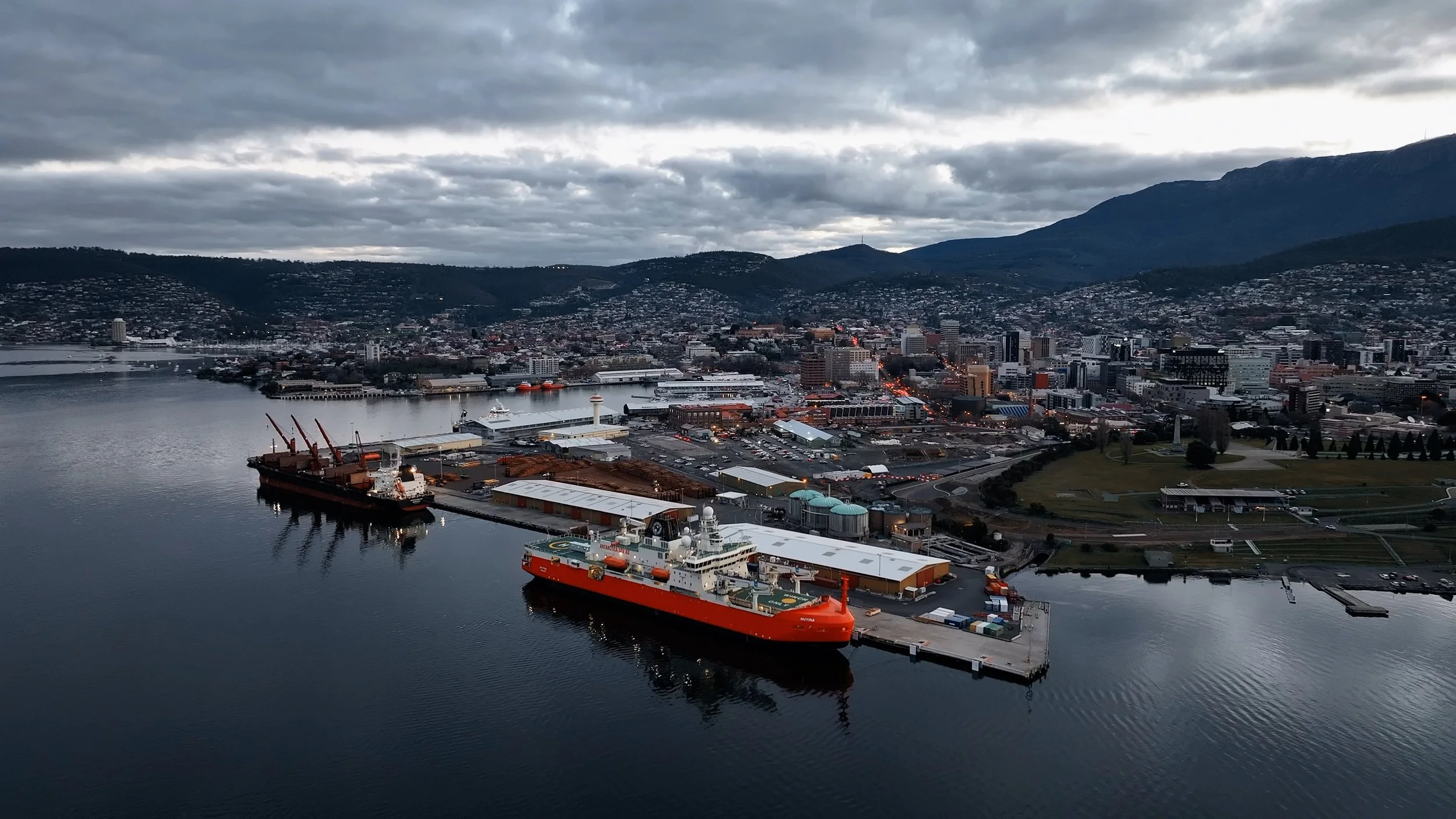 Aerial view of a harbor with ships docked, a cityscape with buildings and streets, and mountains in the background under a cloudy sky.