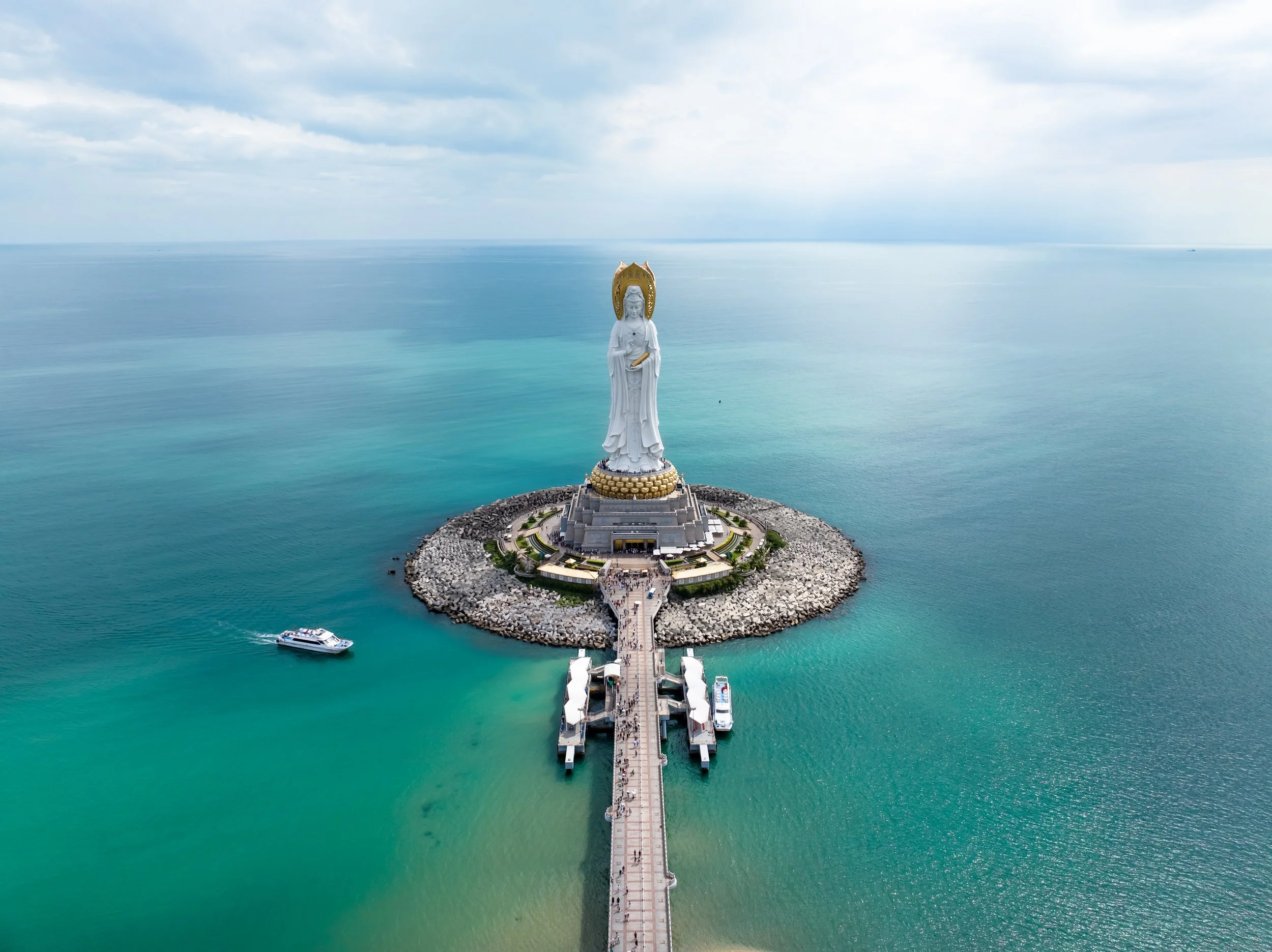 A large white statue of the Goddess of Mercy, Guanyin, stands on a small rocky island in the ocean. The statue is surrounded by a circular platform with a walkway leading to it. A boat is visible near the island, and the ocean and cloudy sky extend i