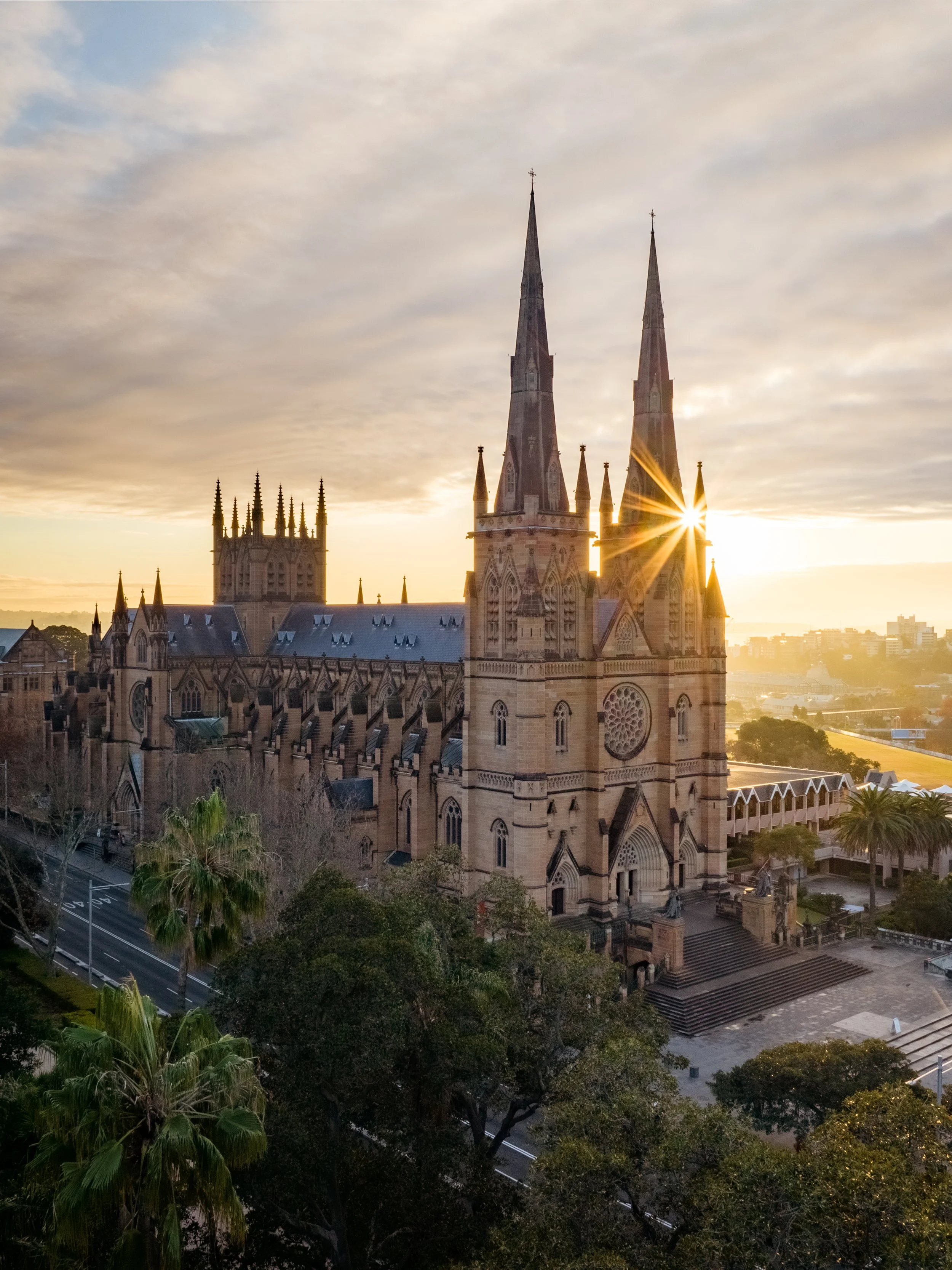 A historic Gothic-style church with tall steeples at sunset, surrounded by trees and a cityscape in the background.