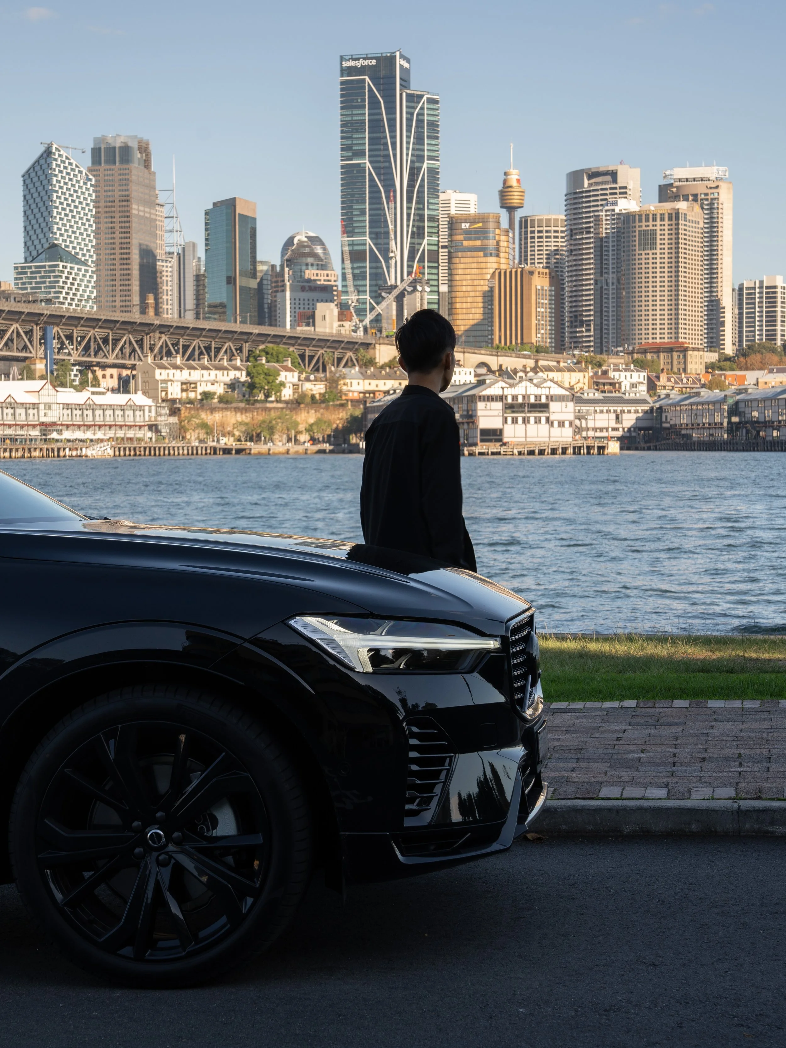 A black luxury car parked near a waterfront with a city skyline in the background, including tall modern skyscrapers and a person standing by the water.