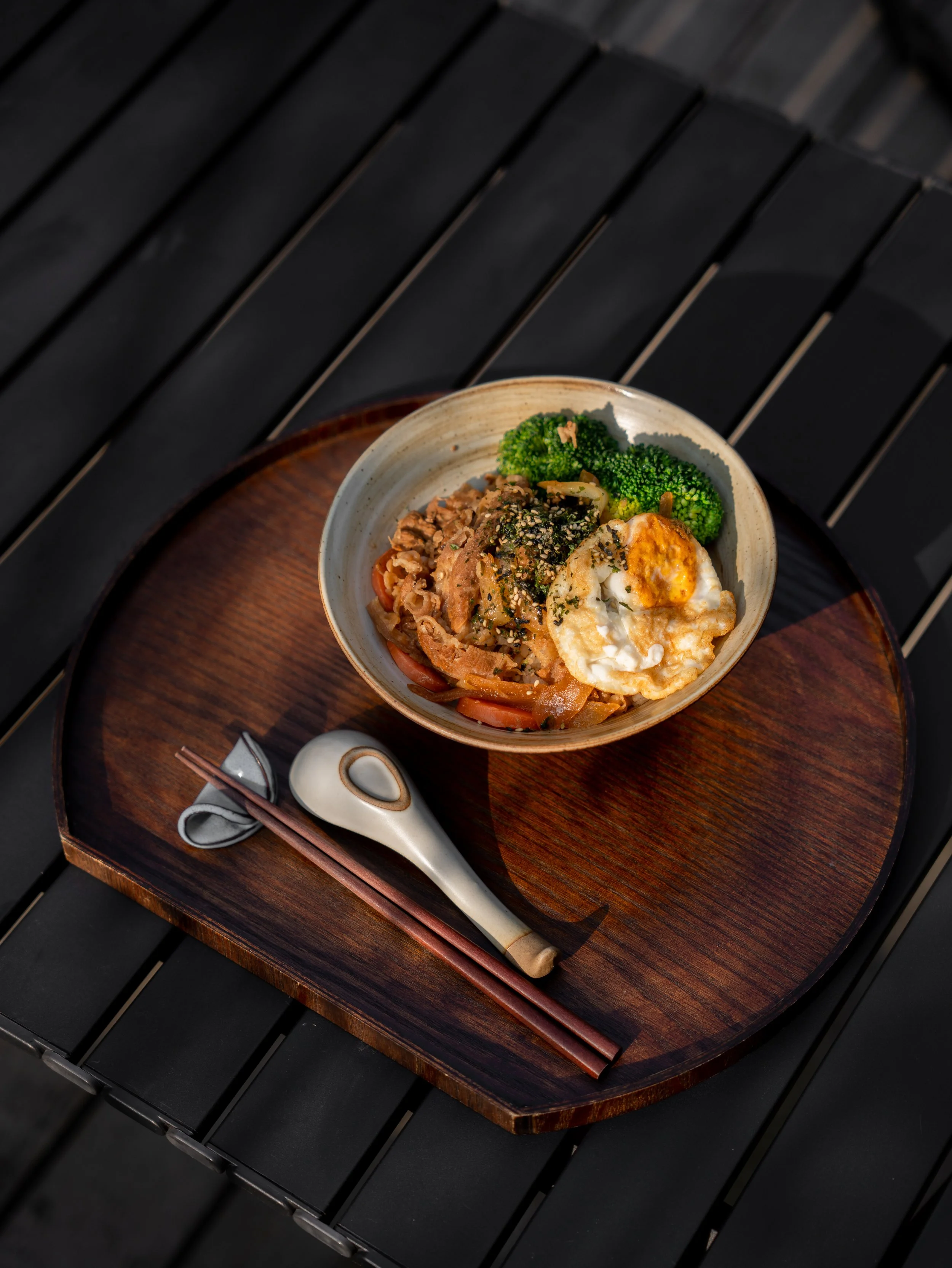 A bowl of Asian cuisine on a wooden tray with chopsticks and a spoon, containing broccoli, rice, and meat.