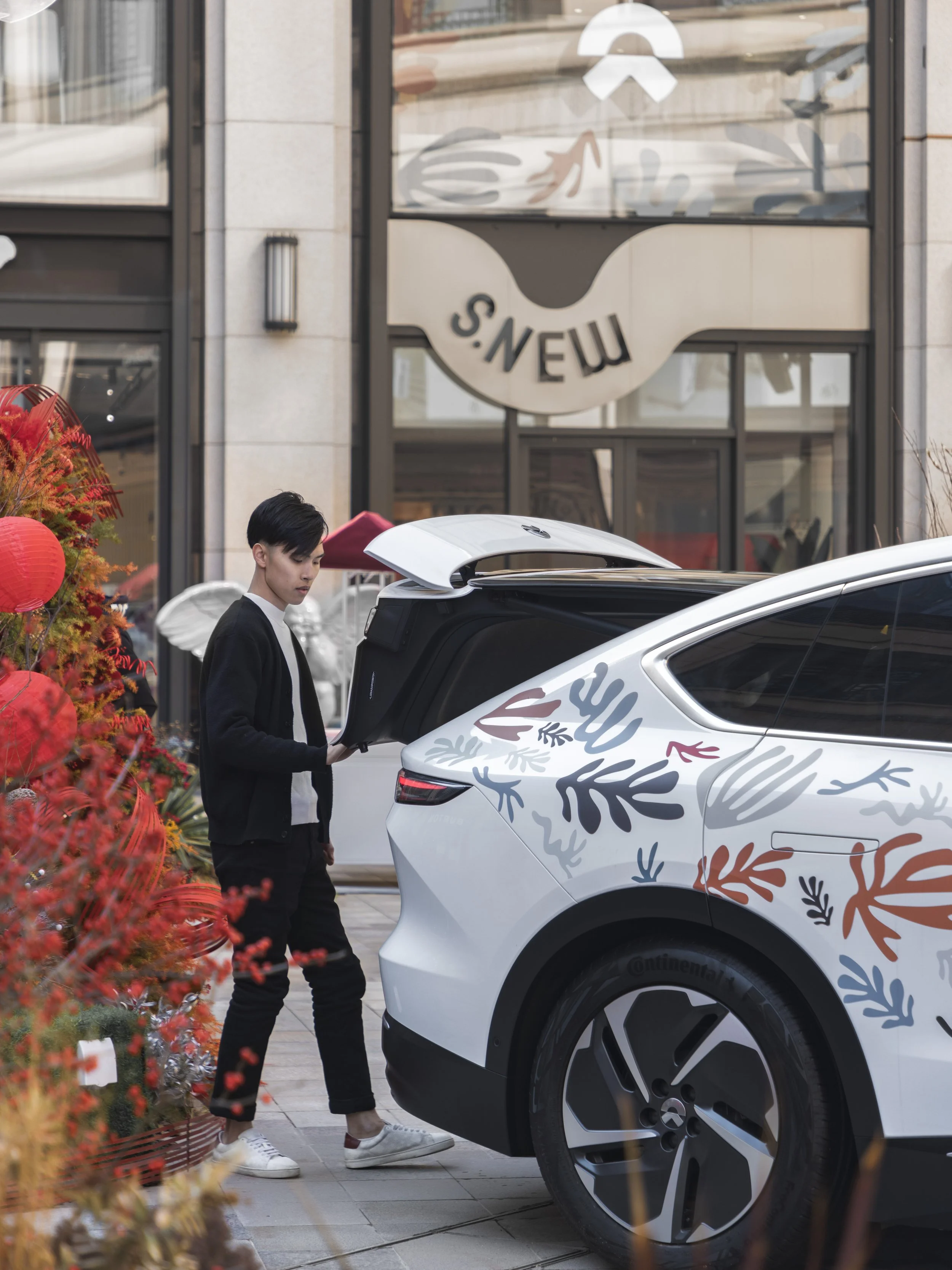 A woman standing behind an electric vehicle with a colorful leaf design on its exterior, opening the trunk in front of a building with a sign that reads 'S. NEW'.