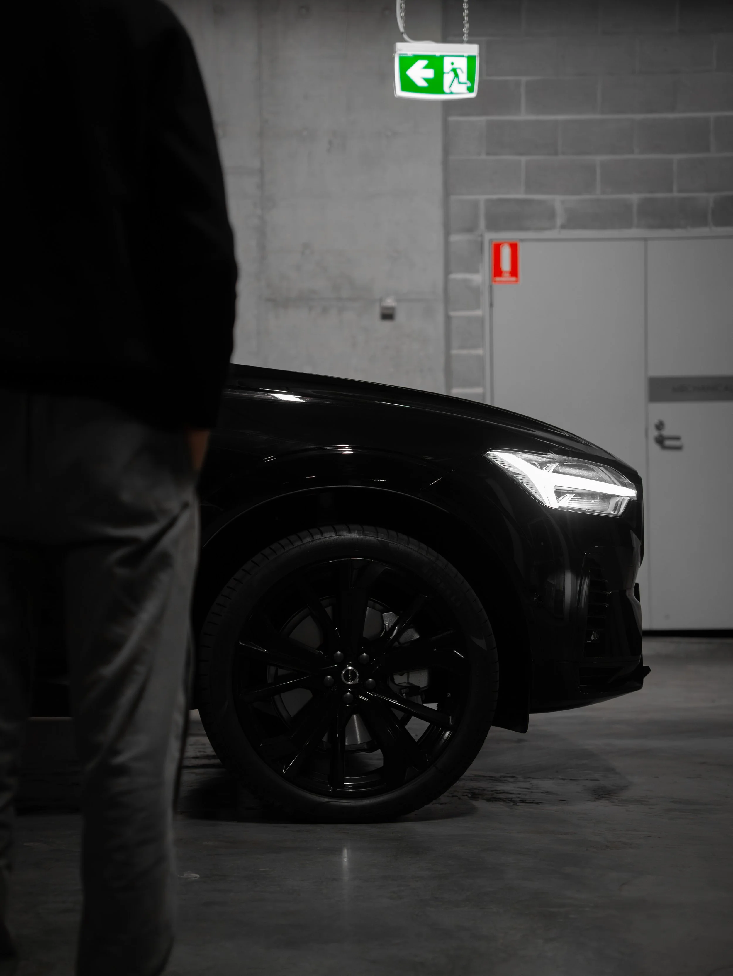 Partially visible black luxury car, possibly an SUV, parked indoors with a concrete and brick wall background. A person wearing camouflage pants and a black jacket is in the foreground, standing next to the car. Above, there is a green illuminated ex