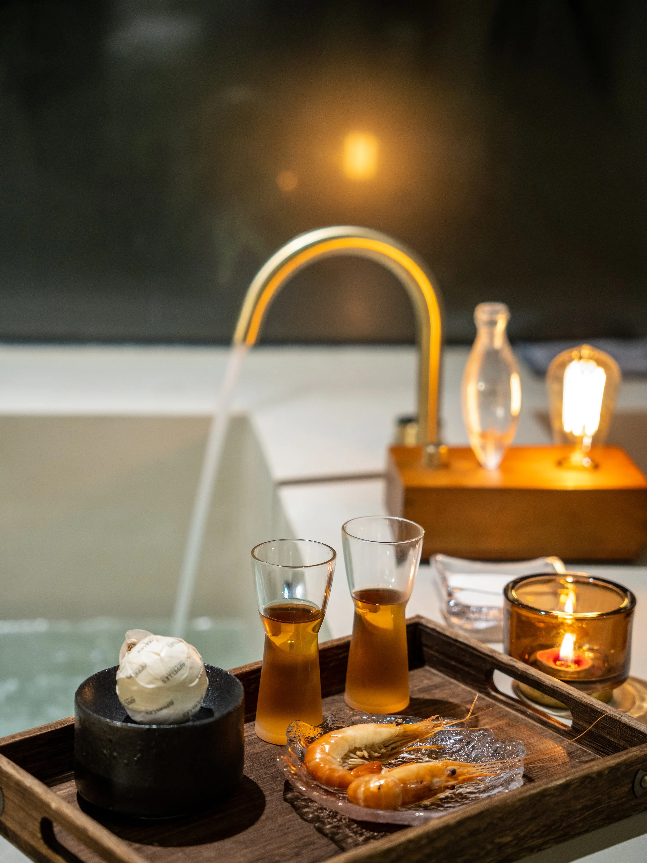 A tray with two glasses of amber-colored beverages, a plate of cooked shrimp on ice, a lit candle in a glass holder, and a small bowl with a wrapped item, with a kitchen sink with a gold faucet in the background.