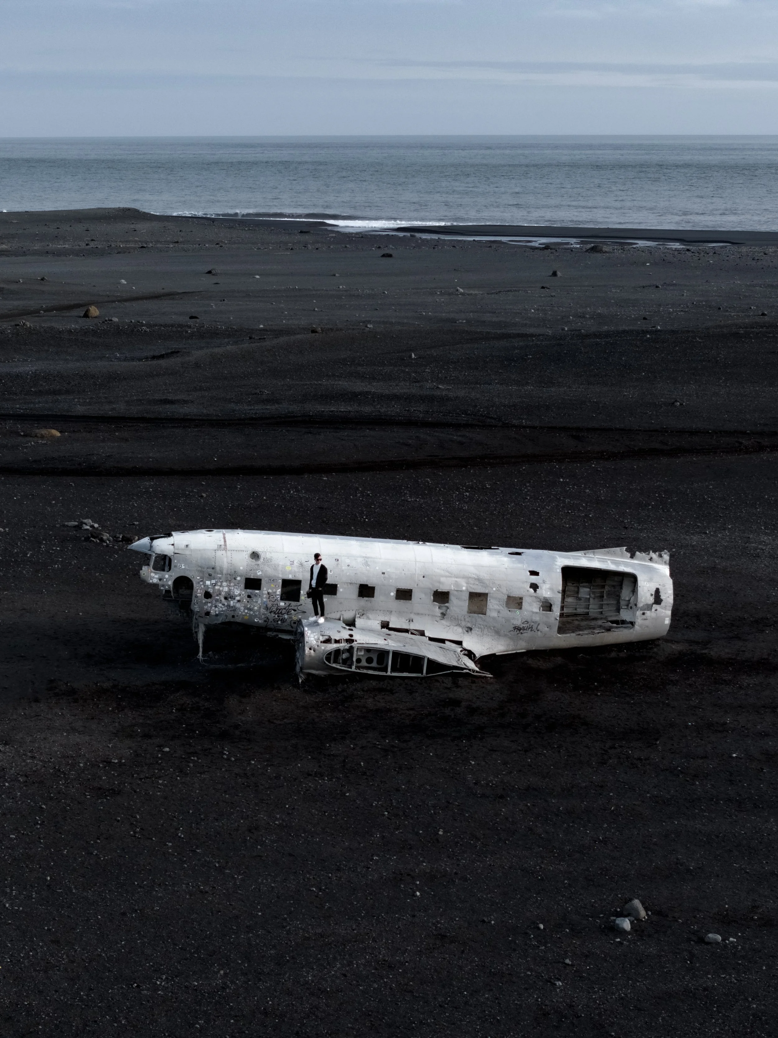 A person stands next to a large, white, wrecked airplane fuselage on a black sandy beach with the ocean and cloudy sky in the background.