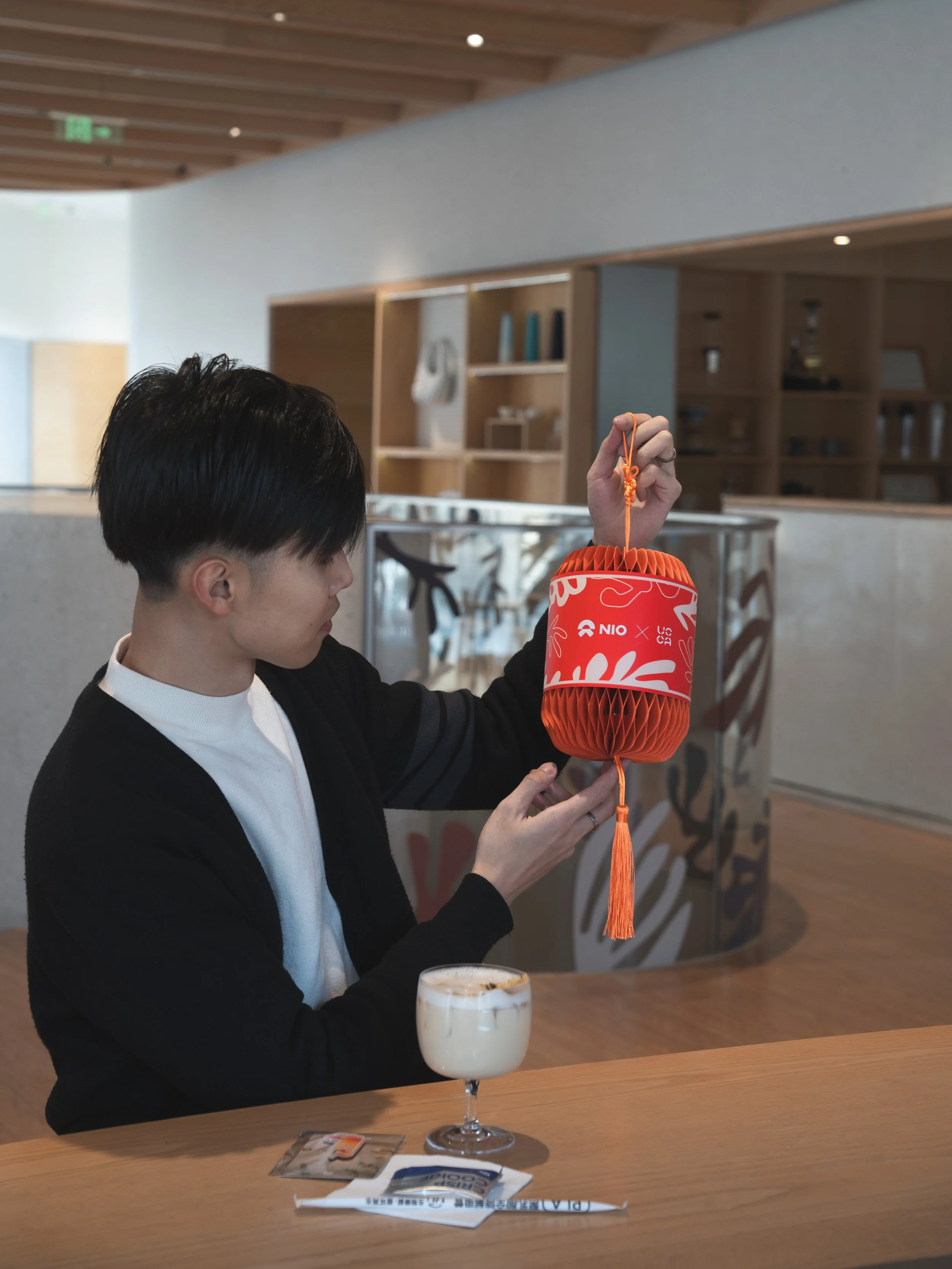 A woman with short black hair wearing a black blazer and white shirt holding an orange paper lantern in a modern cafe or restaurant. There is a latte with foam on the counter in front of her, and a packet of sugar or creamer. The background has woode