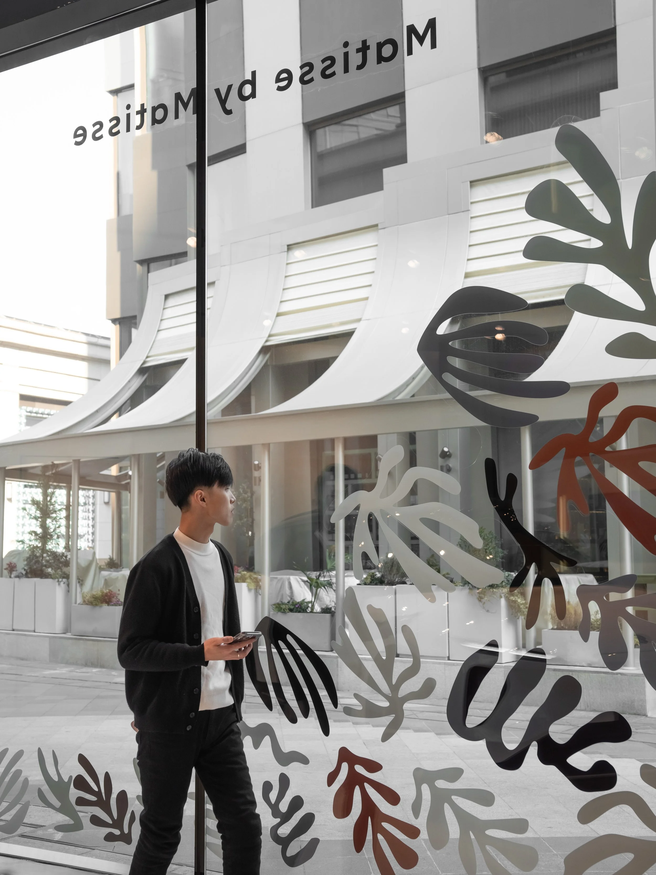 A young man in a black jacket and white T-shirt standing outside a building, looking through a glass window decorated with colorful leaf patterns, holding a smartphone.