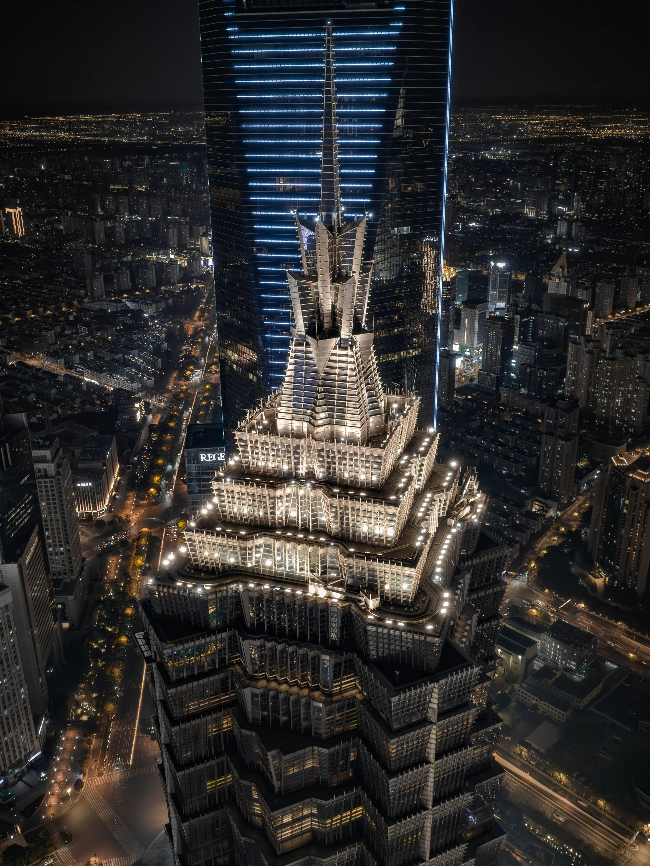 Nighttime aerial view of a tall, illuminated skyscraper with distinctive architectural features, surrounded by city lights and other buildings.