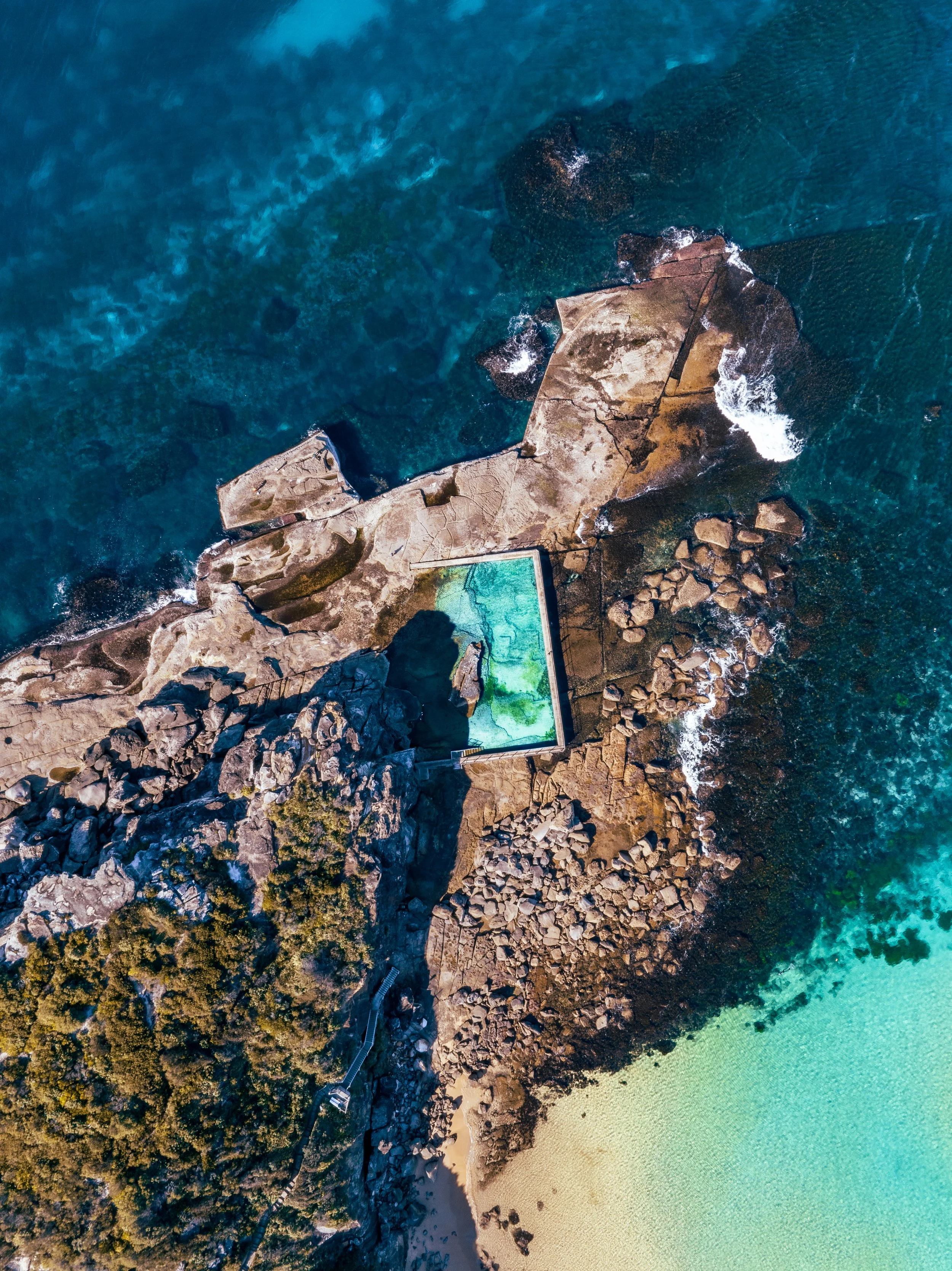 An aerial view of rocky coastline with a swimming pool, dense trees, rocky shoreline, and clear blue-green ocean water.