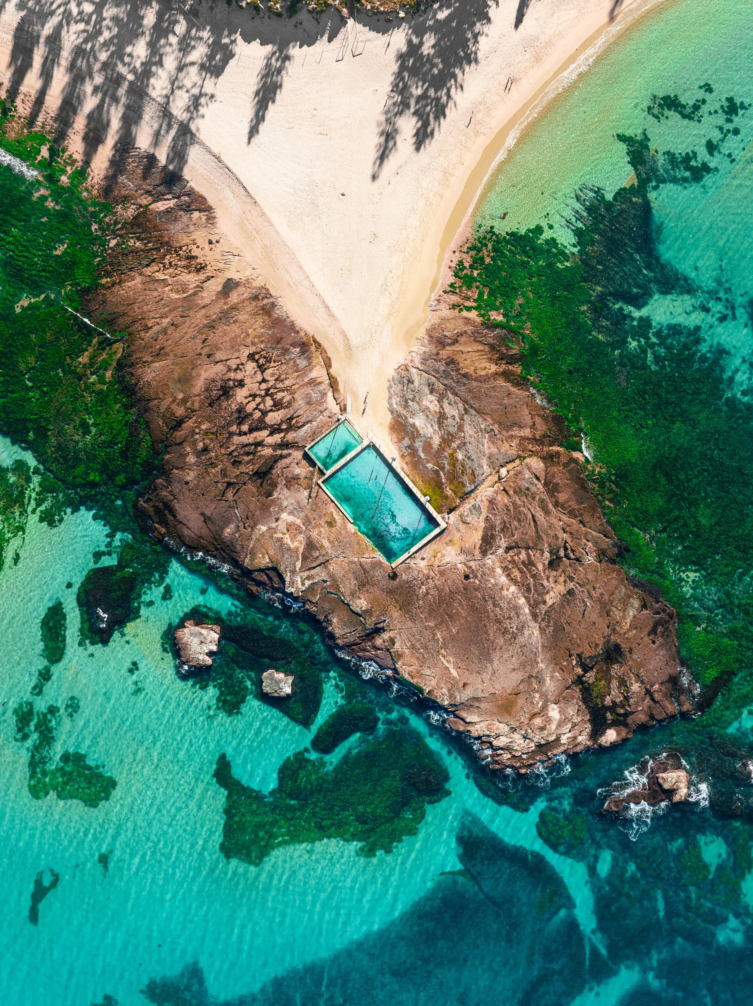 An aerial view of a beach with a sandy shoreline, turquoise waters, green vegetation, and rocky formations. There is a swimming pool on the rocky area near the water.