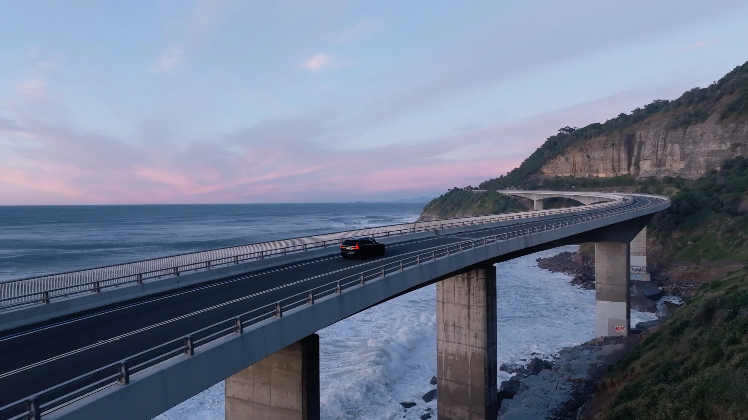 A black SUV driving on a coastal bridge with a rocky hillside and the ocean in the background during sunset.