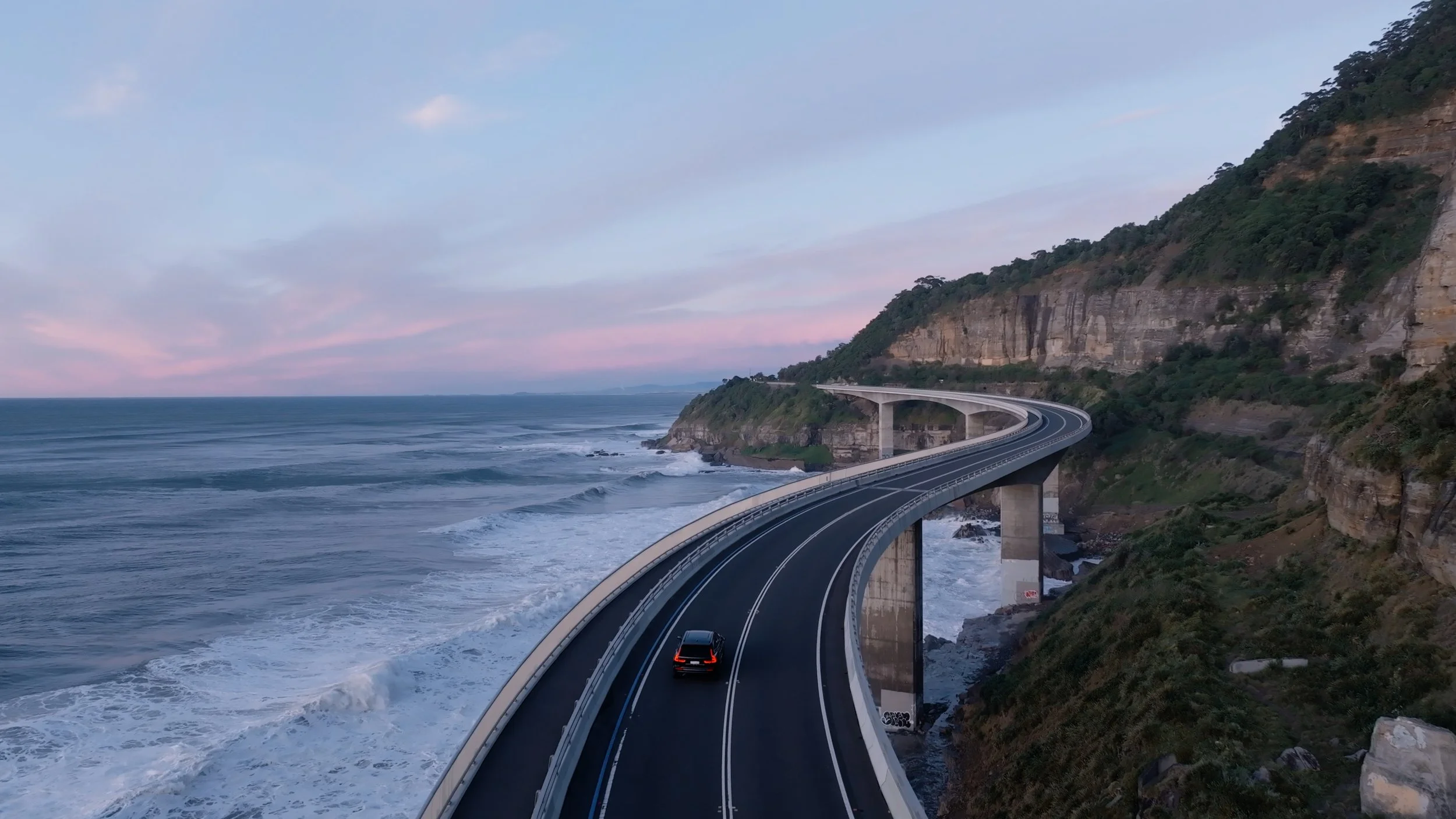 A coastal highway curves along a steep cliffside with ocean waves crashing below. The road is elevated on concrete supports and surrounded by rocky terrain and green vegetation.