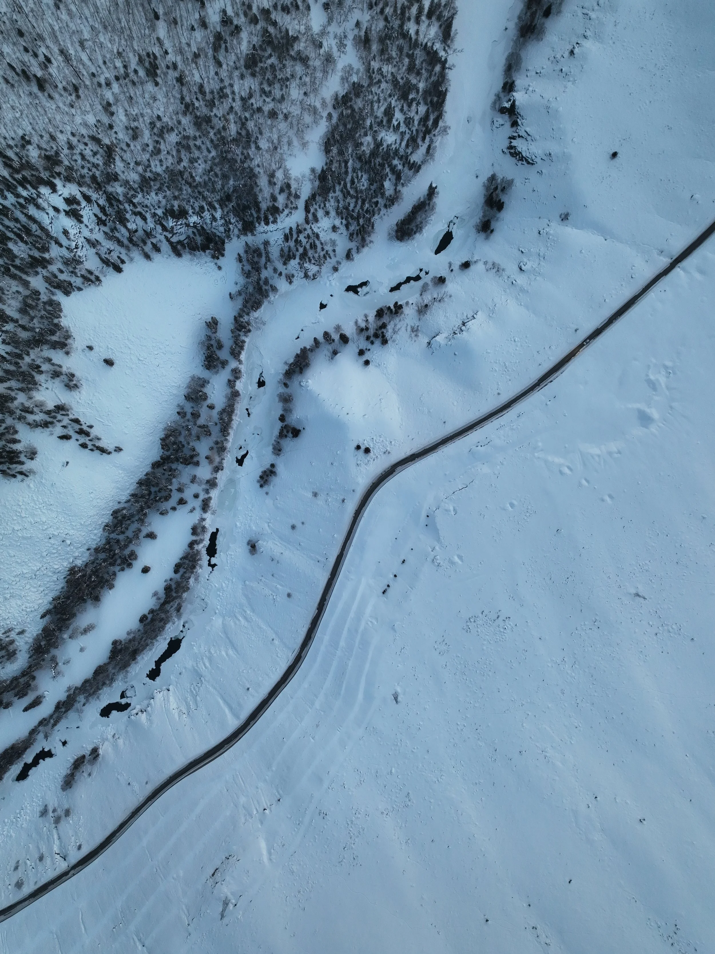 A snowy landscape with a narrow river or stream running through snow-covered terrain. A winding road is visible on the snow, and there is a dense forest of trees on one side of the stream.