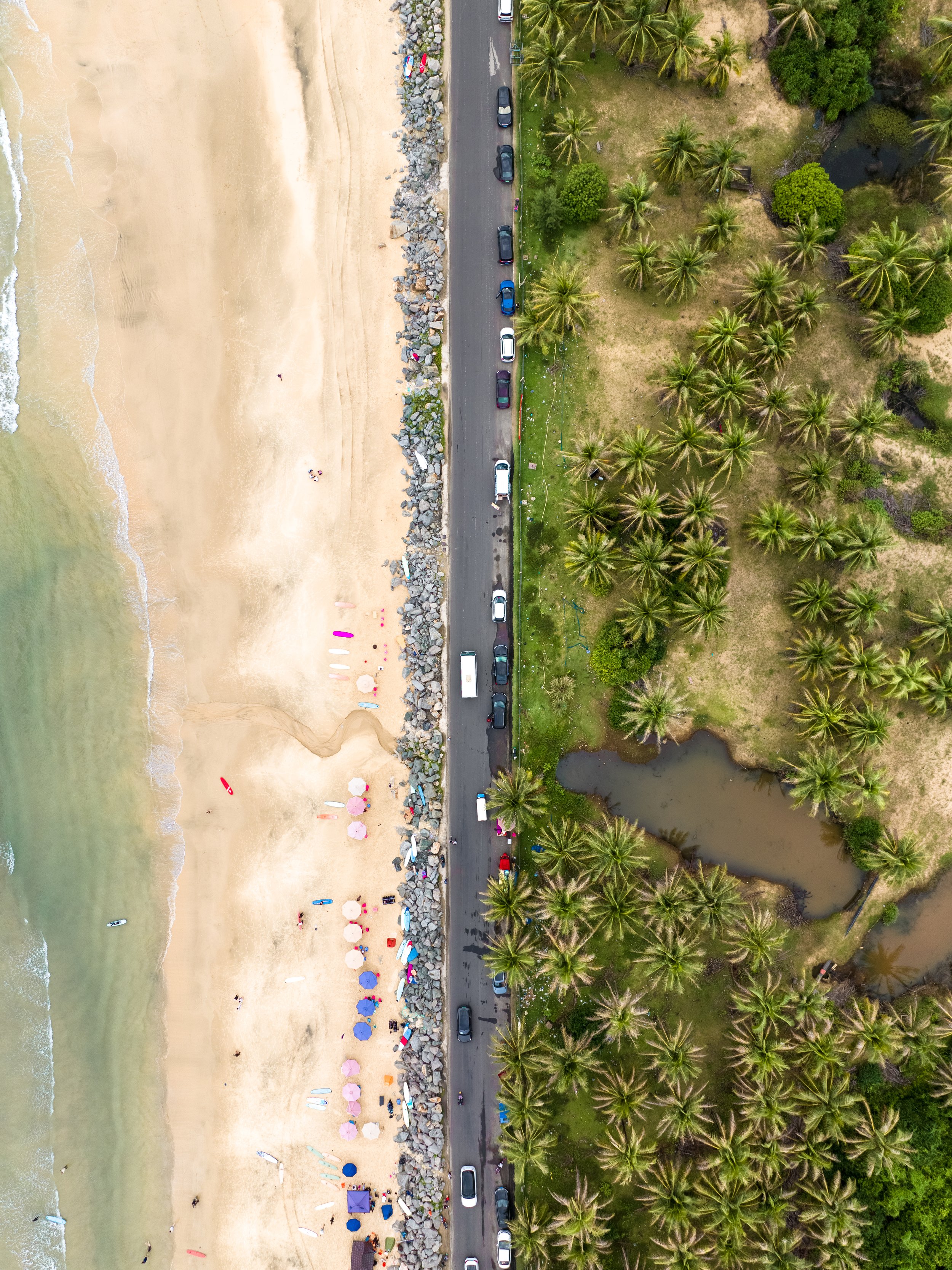 An aerial view of a beach with colorful umbrellas, lounge chairs, and people near the shoreline. A rocky breakwater runs parallel to the beach, separating the sandy area from the road and a row of cars. To the right of the road, there are lush green 