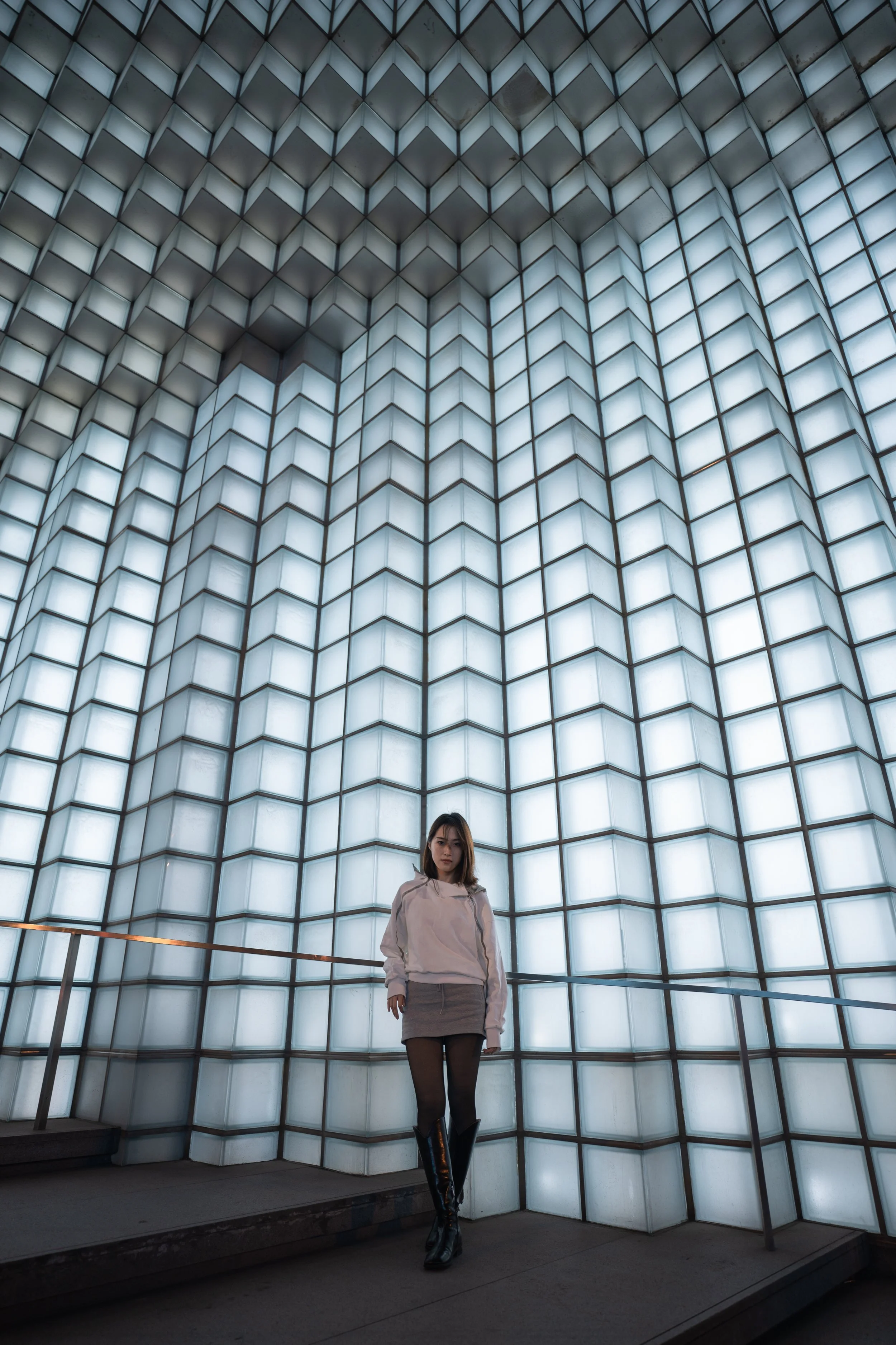 A young woman in a light-colored jacket, black tights, and knee-high black boots standing in front of a wall of illuminated glass blocks at night.