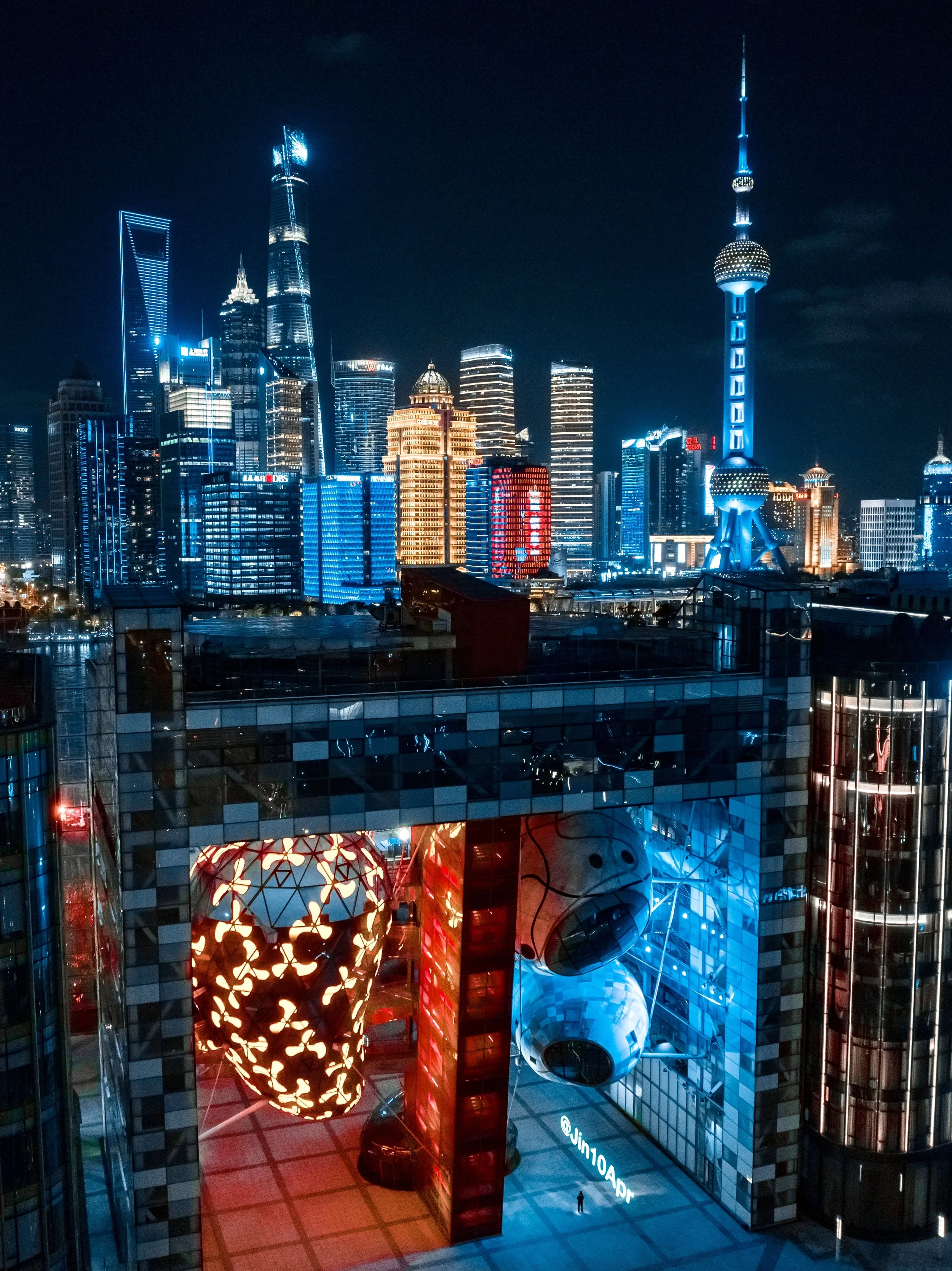 Night view of Shanghai skyline with illuminated skyscrapers, including the Oriental Pearl Tower and Shanghai World Financial Center, seen from an indoor area with colorful illuminated architecture and a person walking below.