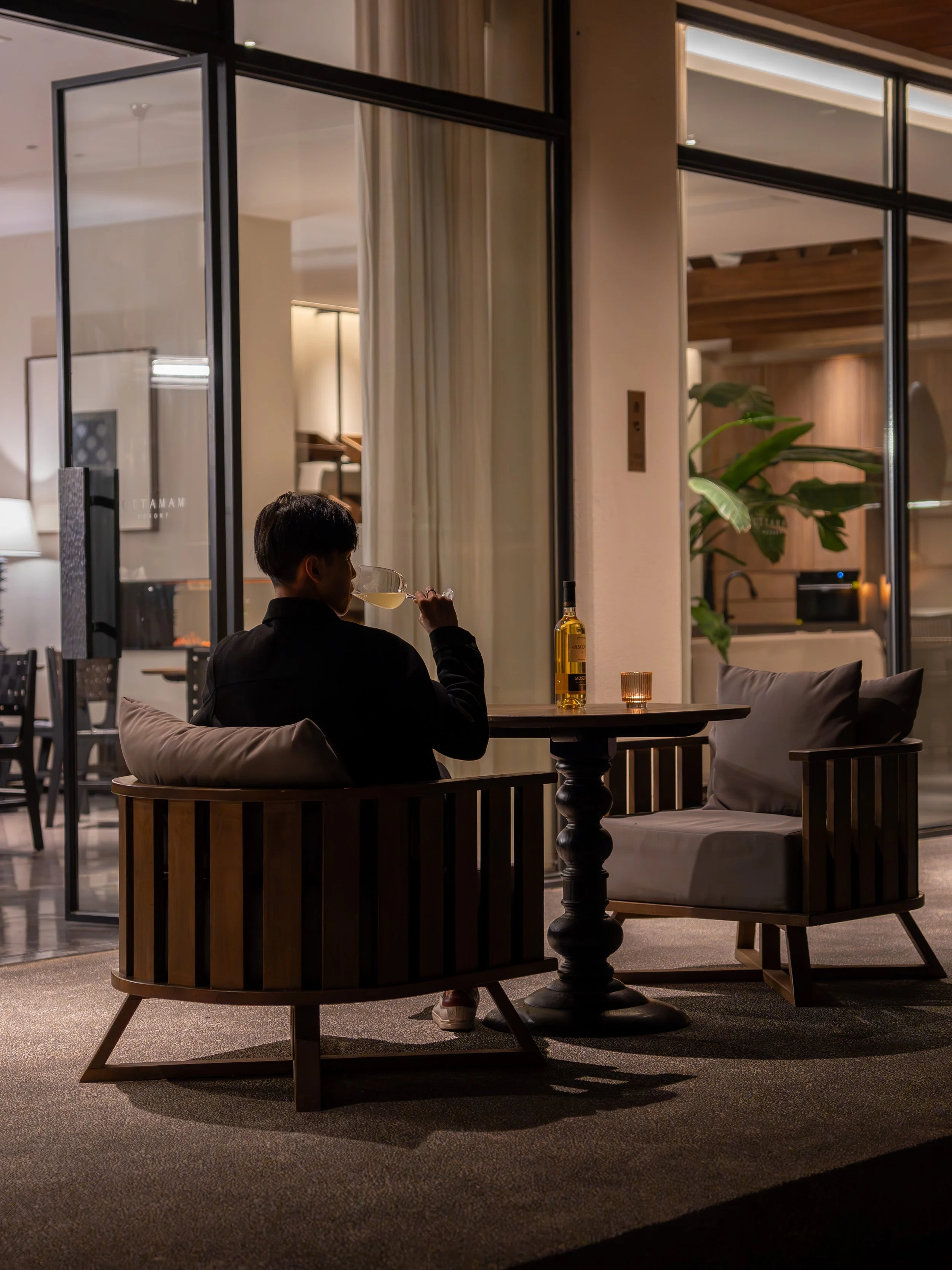 A person sitting in a modern, well-lit indoor space at night, drinking a glass of white wine or champagne, with a bottle of alcohol, candle, and comfortable furniture nearby, surrounded by large glass windows and greenery.