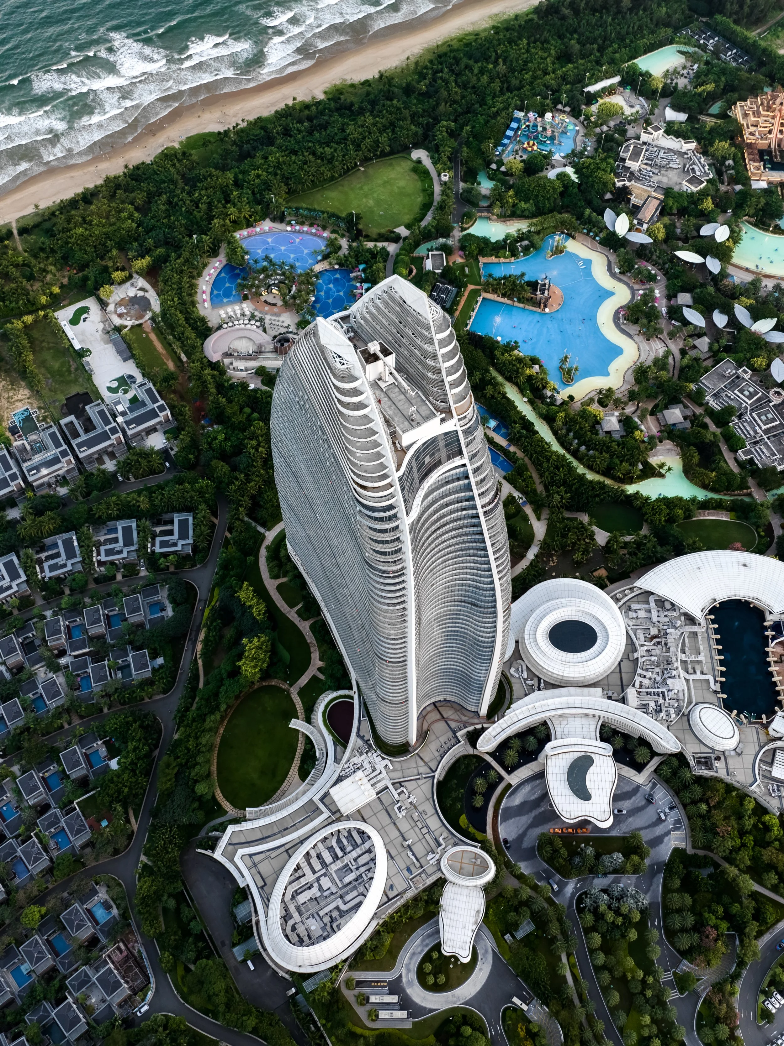 Aerial view of a beachfront resort with a tall modern hotel building, multiple pools, and surrounding greenery along the coast.