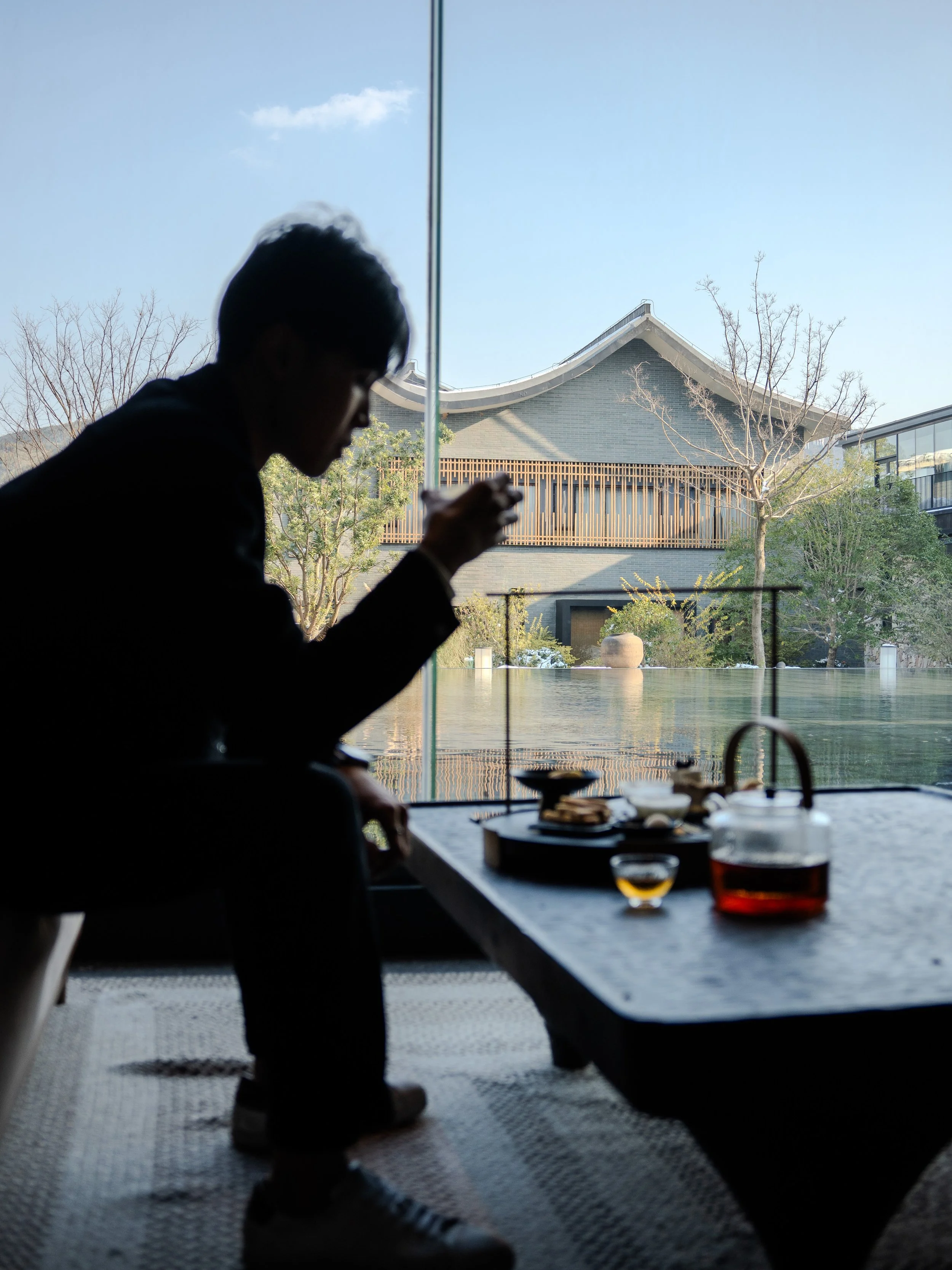 Silhouette of a person sitting at a table with food, seen in front of a large window overlooking a building and trees outside.