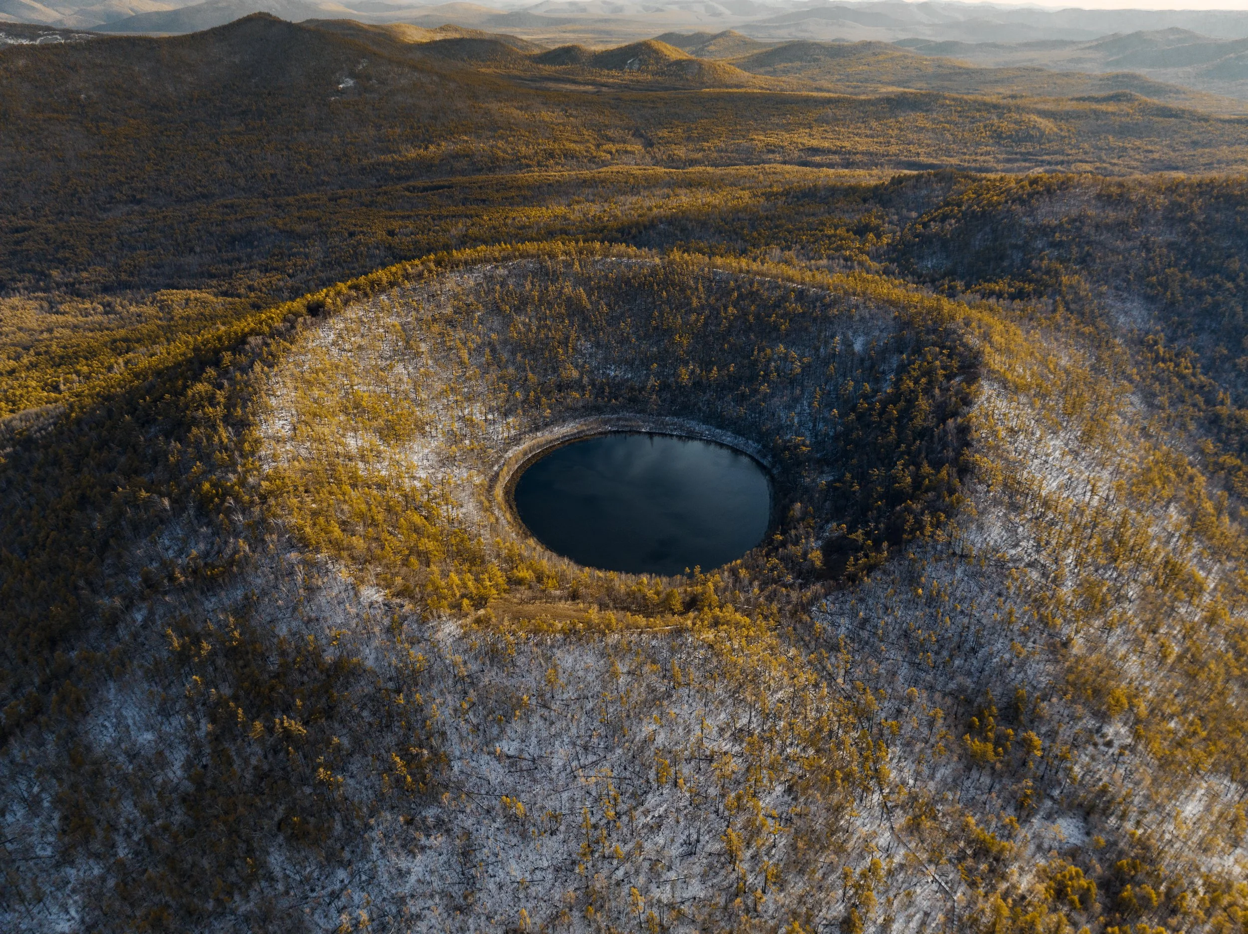 Aerial view of a mountain landscape with a dark round lake at the center, surrounded by forested hills with some patches of snow, during winter.