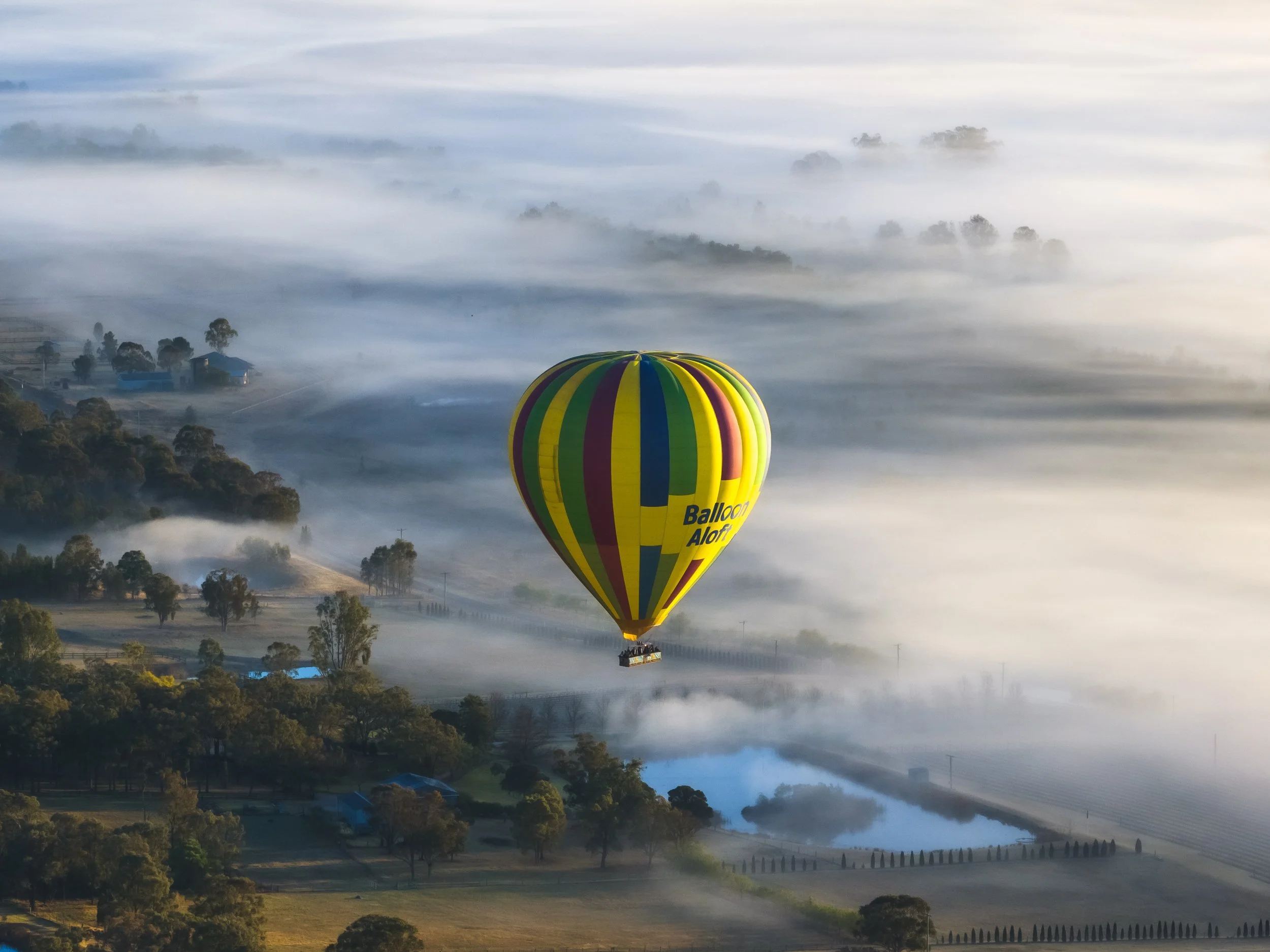 A colorful hot air balloon floating above a foggy, rural landscape during dawn or early morning.
