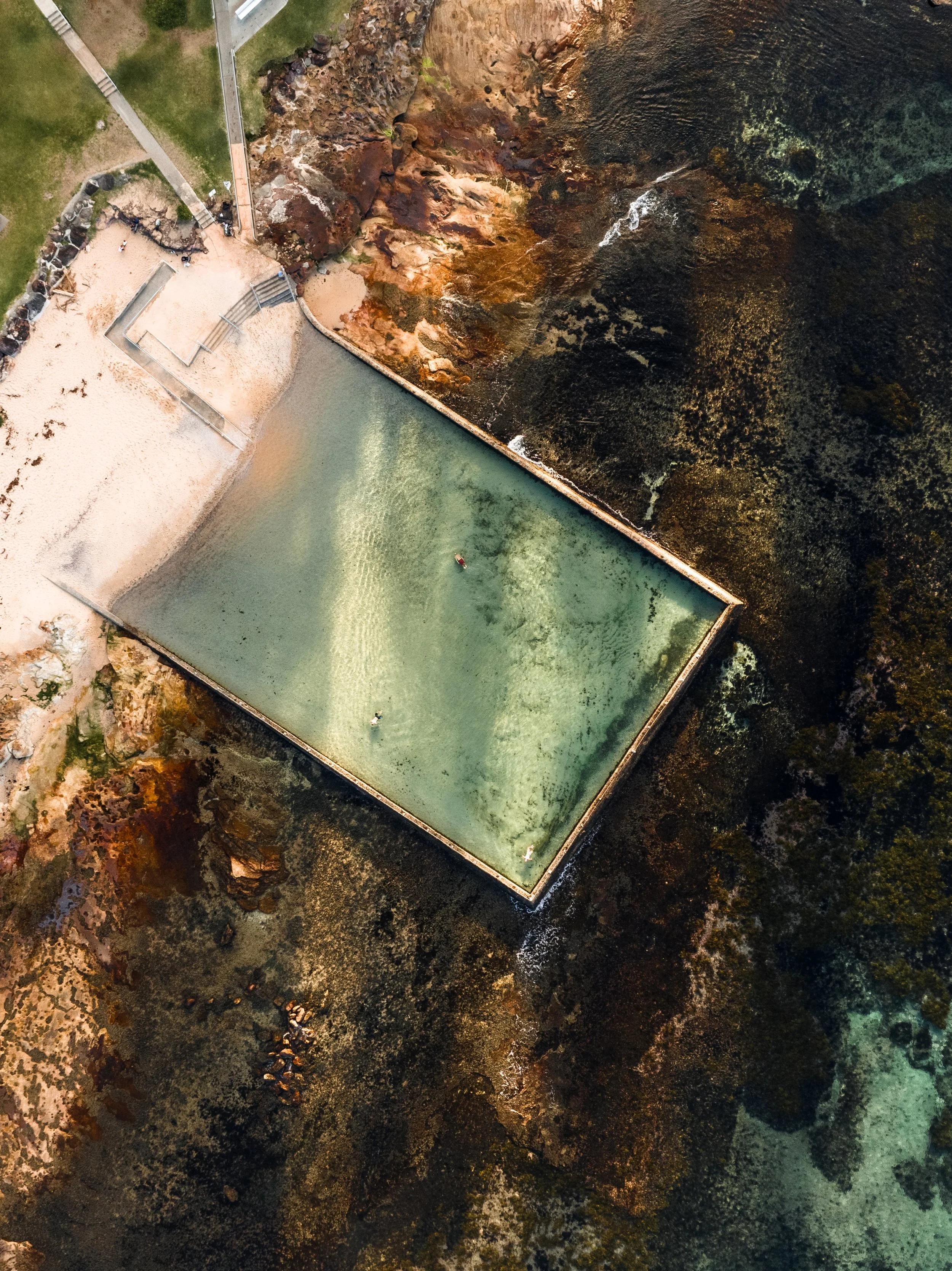 An aerial view of a saltwater swimming pool surrounded by rust-colored rocks and dark water, with a small boat in the pool and a sandy area adjacent to it.