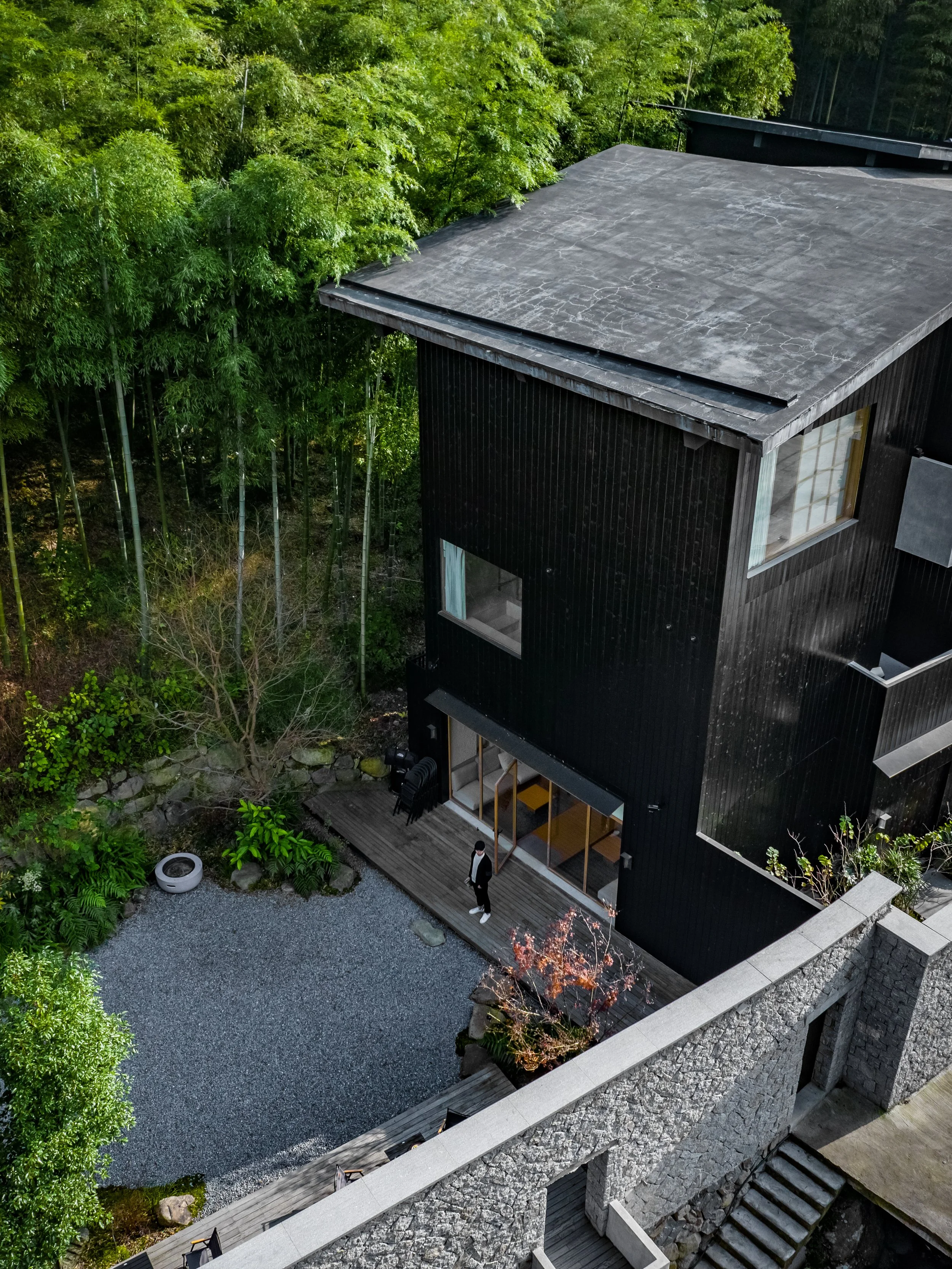 A modern black house with a flat roof, set amidst green trees and vegetation. The house has large windows and a small outdoor deck with a person standing on it. The yard features gravel, plants, and a stone retaining wall.