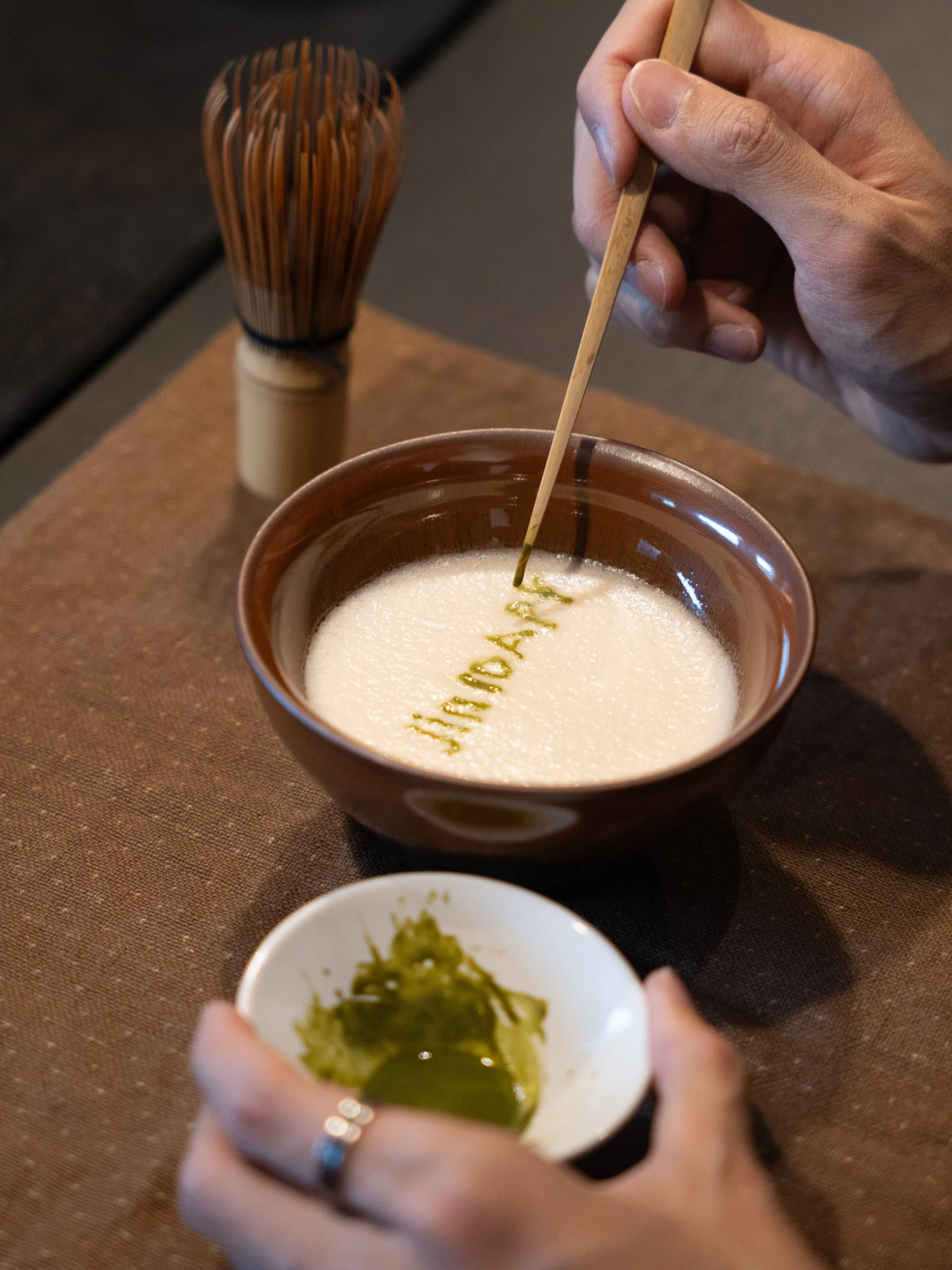 A person holds a small bowl with green wasabi in front of a larger bowl of foamy, frothy beverage with the word 'HAMACHI' written on it. A wooden utensil stirs the drink, and an ornate wooden soy sauce dispenser is in the background.