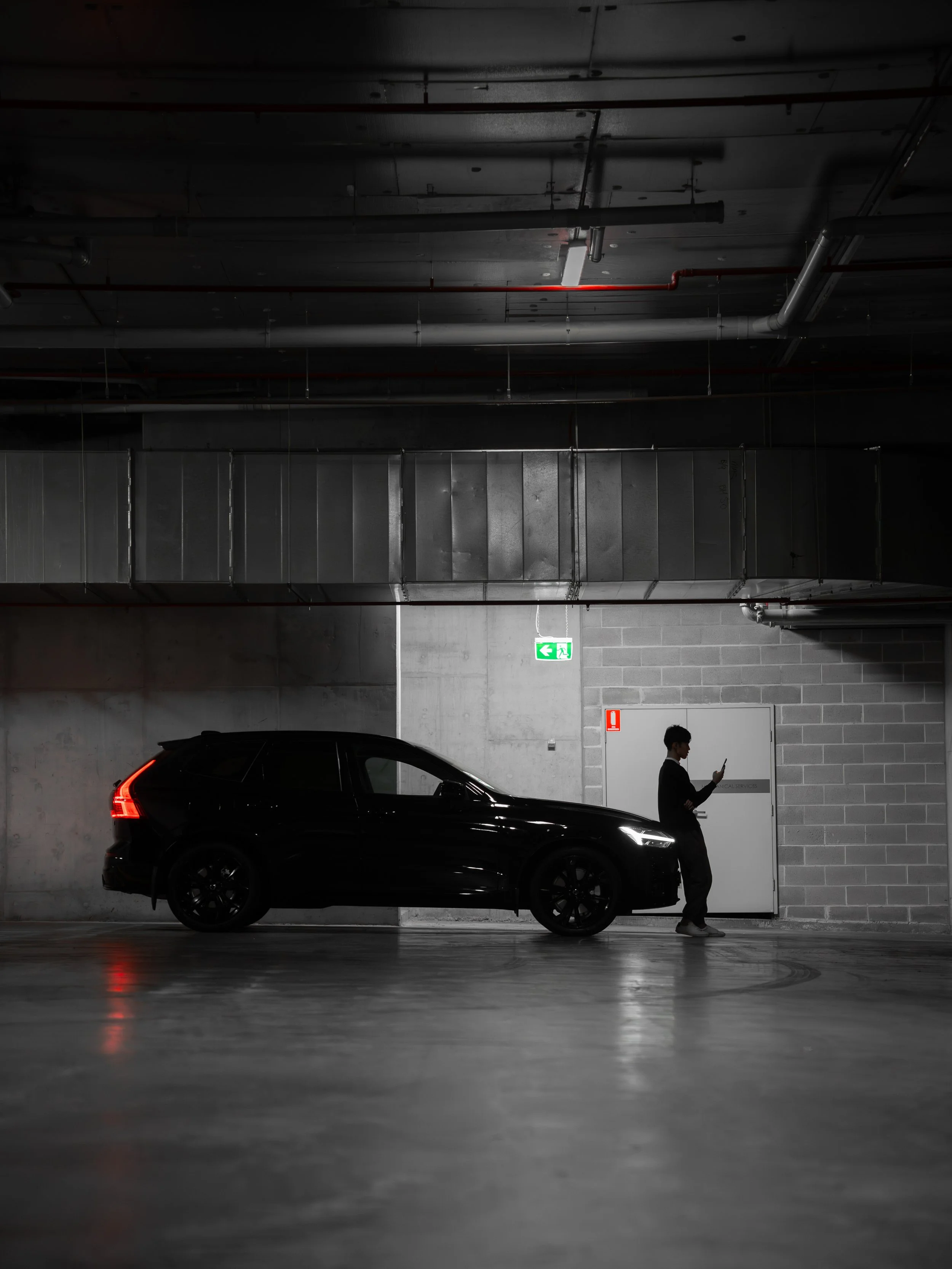 A black car parked in an underground parking garage with a person walking past while looking at their phone.