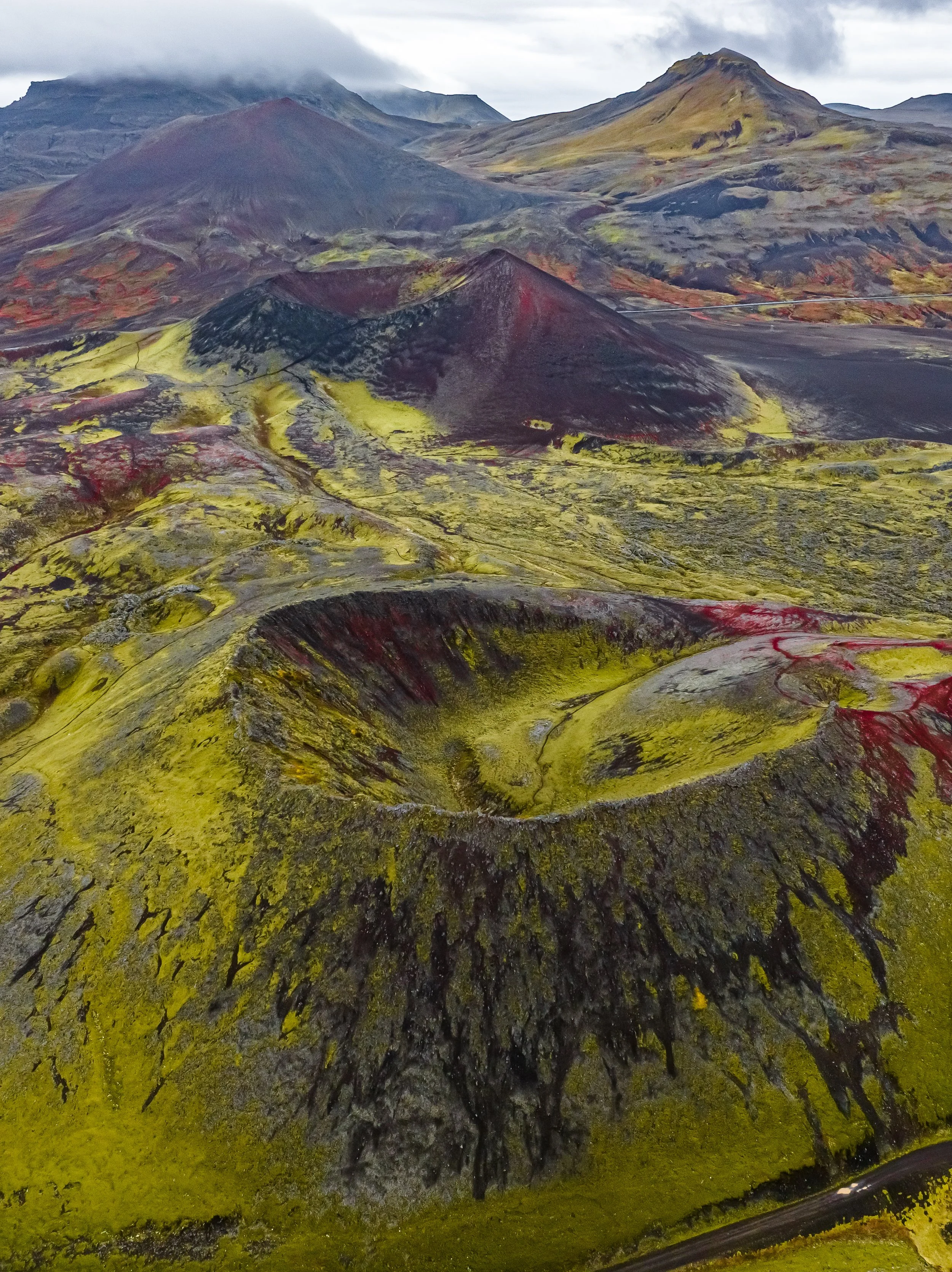 Volcanic landscape with colorful moss and lava flows, mountains in the background, and a cloudy sky.