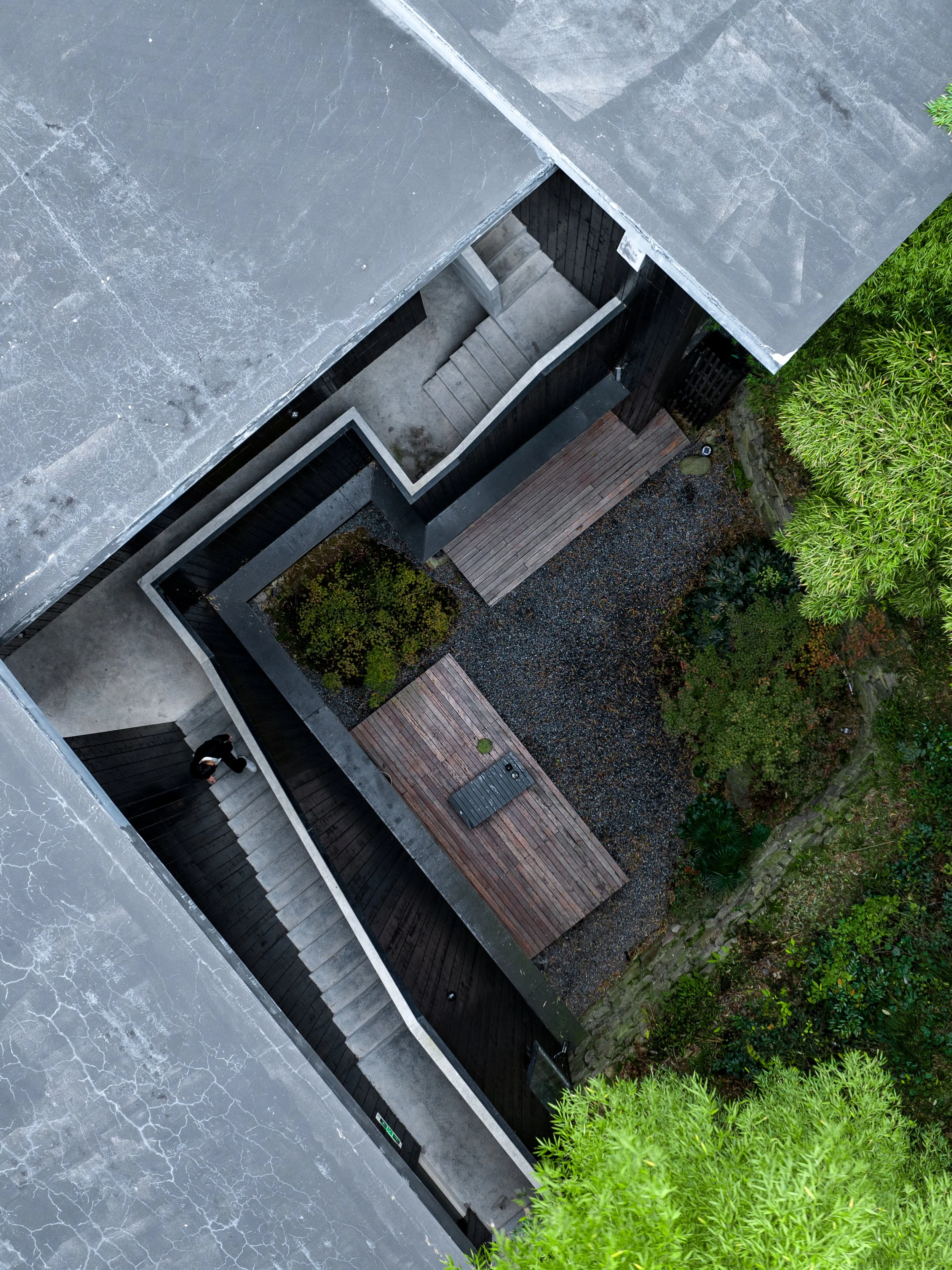Aerial view of a modern building with black exterior walls, concrete stairs, a wooden deck, and surrounding landscaping with green bushes and gravel ground, viewed from above.