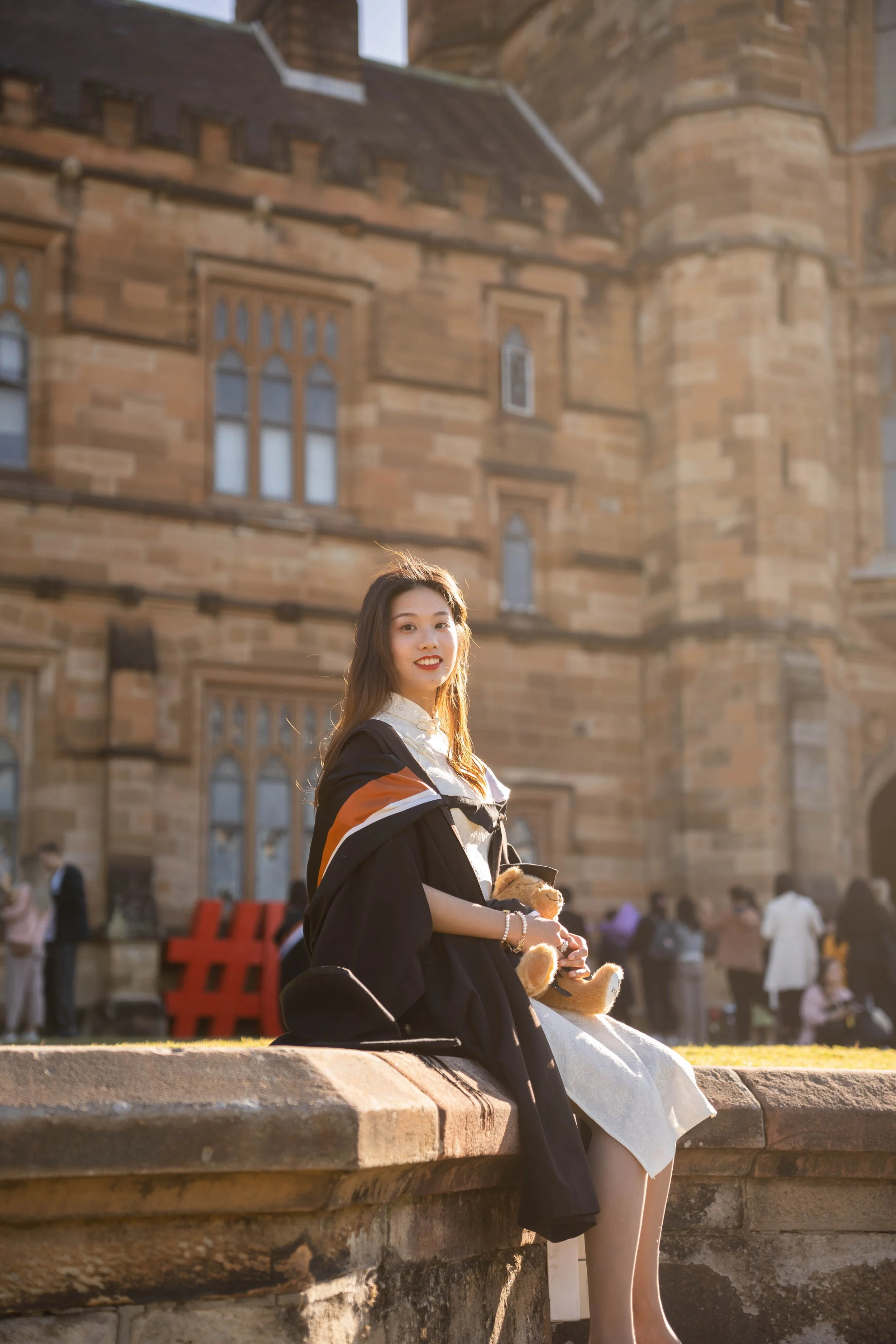 A young woman in a graduation gown sitting on a stone ledge holding a teddy bear, with a historic brick building in the background.