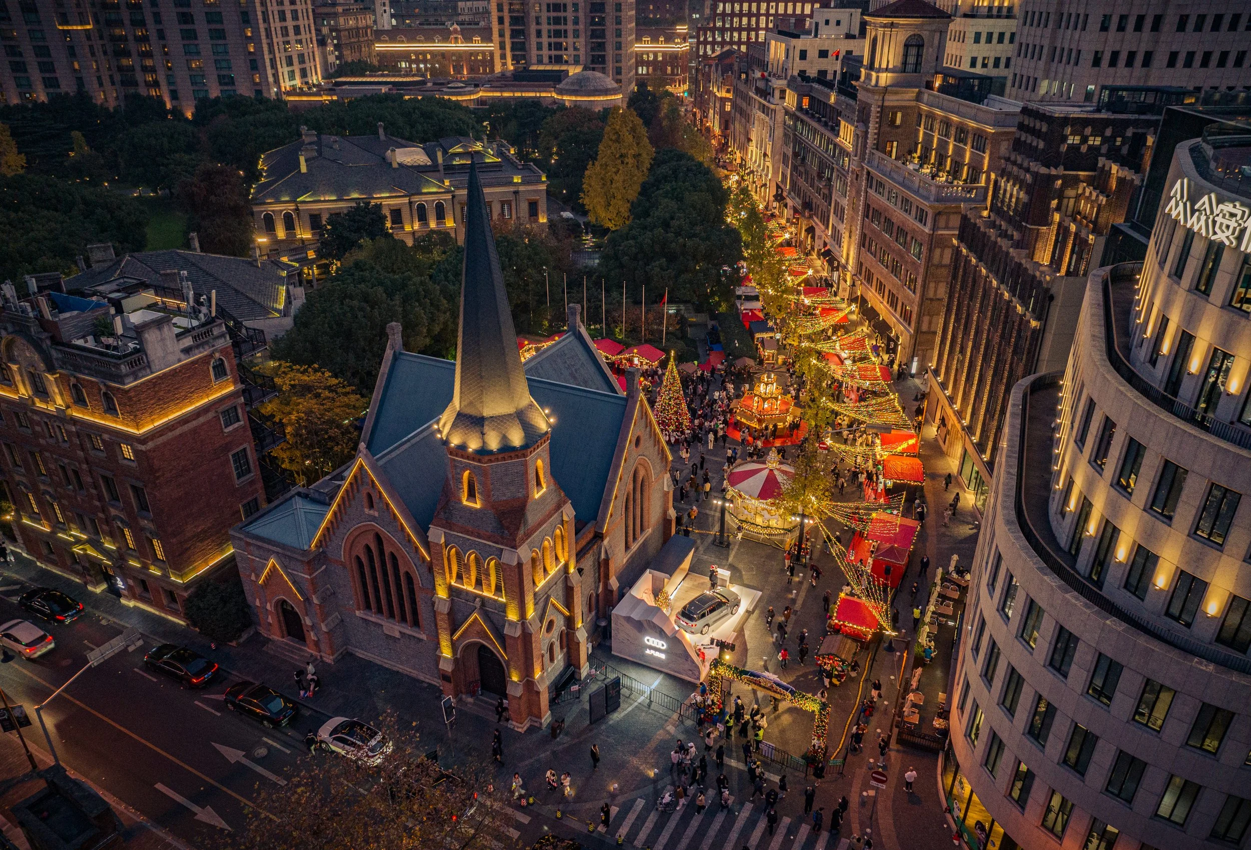 Night view of a city street decorated for the holidays with a festive carnival, Christmas trees, colorful lights, and a crowd of people.